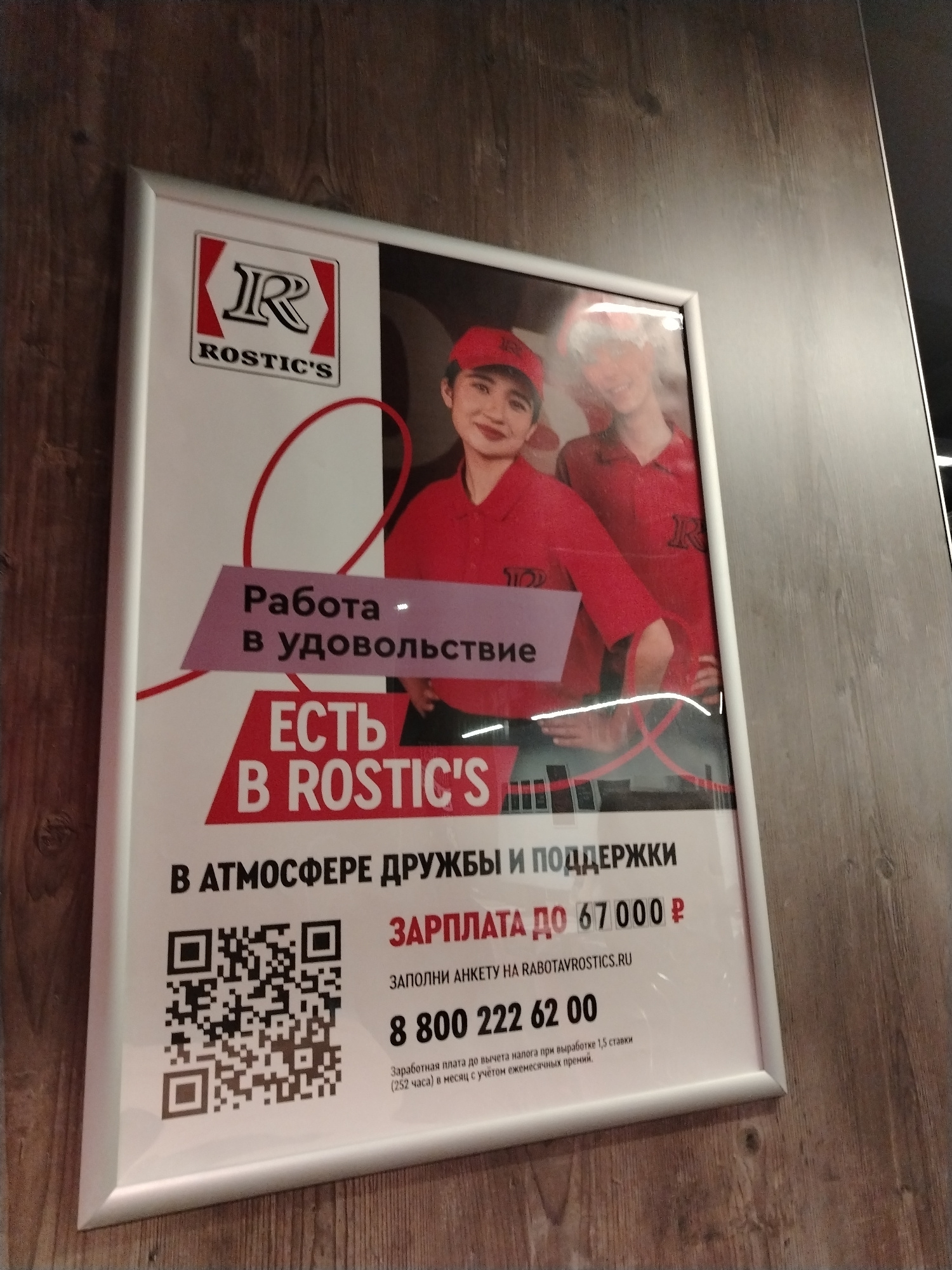 The image shows a framed poster with the logo "Rostic's" and text in Russian. The poster is hanging on a wooden wall, likely in a public space in Yaroslavl, Russia. The poster features two young women in red uniforms, smiling. Text on the poster advertises "Work with pleasure" and "There is at Rostic's" with an emphasis on "atmosphere of friendship and support." It also mentions a salary up to 67,000 rubles and provides a QR code and a phone number for job applications. The overall impression is a recruitment advertisement for employment at a fast-food establishment.