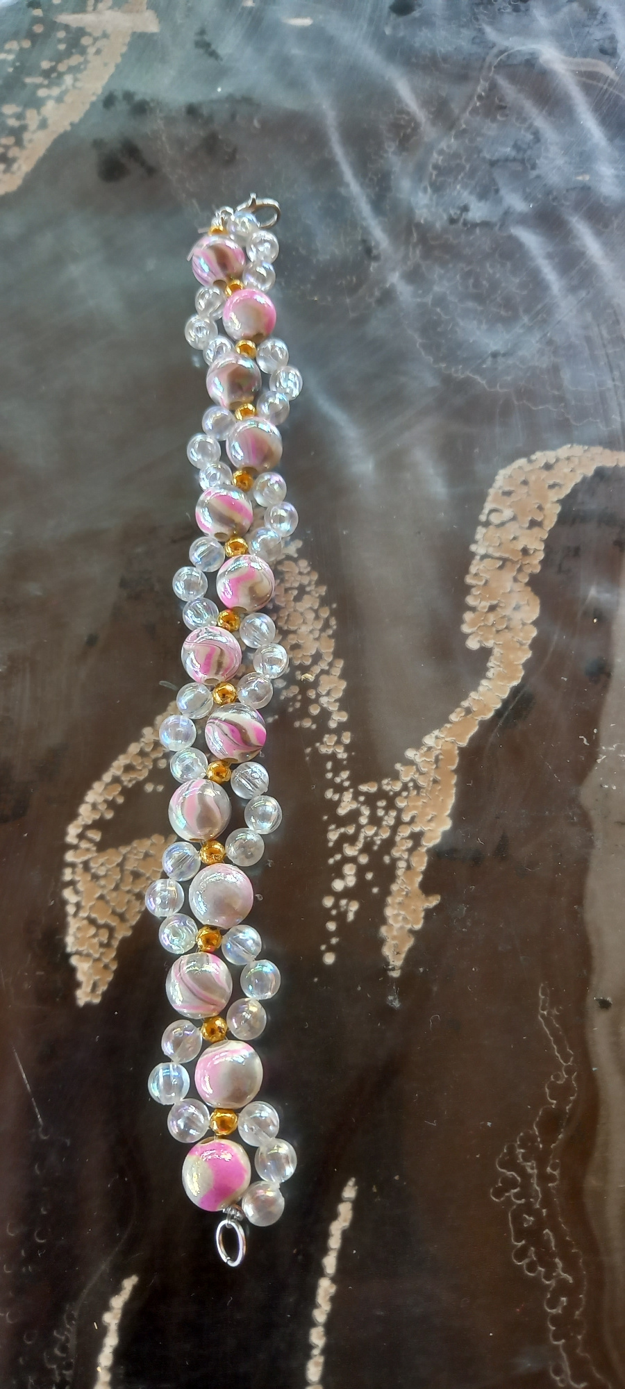 The image shows a beaded bracelet lying on a dark, patterned surface. The bracelet is made of alternating clear, iridescent, and pink marbled beads, with small gold beads in between. The background appears to be a dark, glossy material with swirling patterns of lighter color, possibly a table or countertop. The bracelet is positioned vertically on the left side of the frame, with its clasp visible at the top. The image is taken from above, looking down at the bracelet. There is no visible indication of Iskra, Bulgaria, in the image.