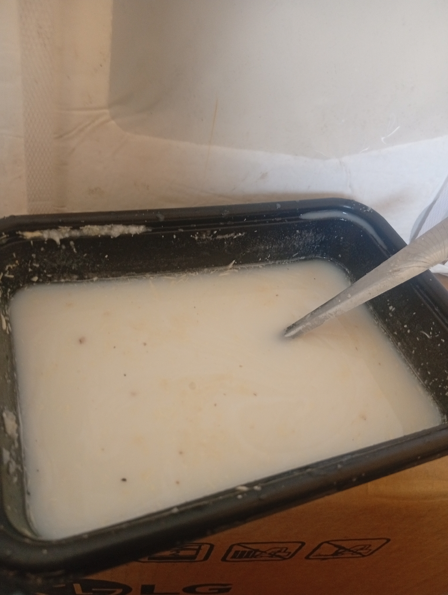 A close-up, overhead shot shows a black rectangular plastic container filled with a milky white liquid, possibly soup or porridge. A silver, pointed utensil, resembling a small trowel or scoop, is submerged in the liquid, with its tip angled downwards. The liquid has small, dark specks suspended within it. The container's rim is speckled with dried, white residue. The background consists of a textured, light-colored surface, possibly a tablecloth or a wall, and a portion of a brown, possibly wooden surface with embossed symbols, one of which appears to be a stylized "LG" logo. There are no people visible in the image. The scene is indoors, with neutral lighting. The location in Bukuru, Nigeria, is not evident from the visual information provided.