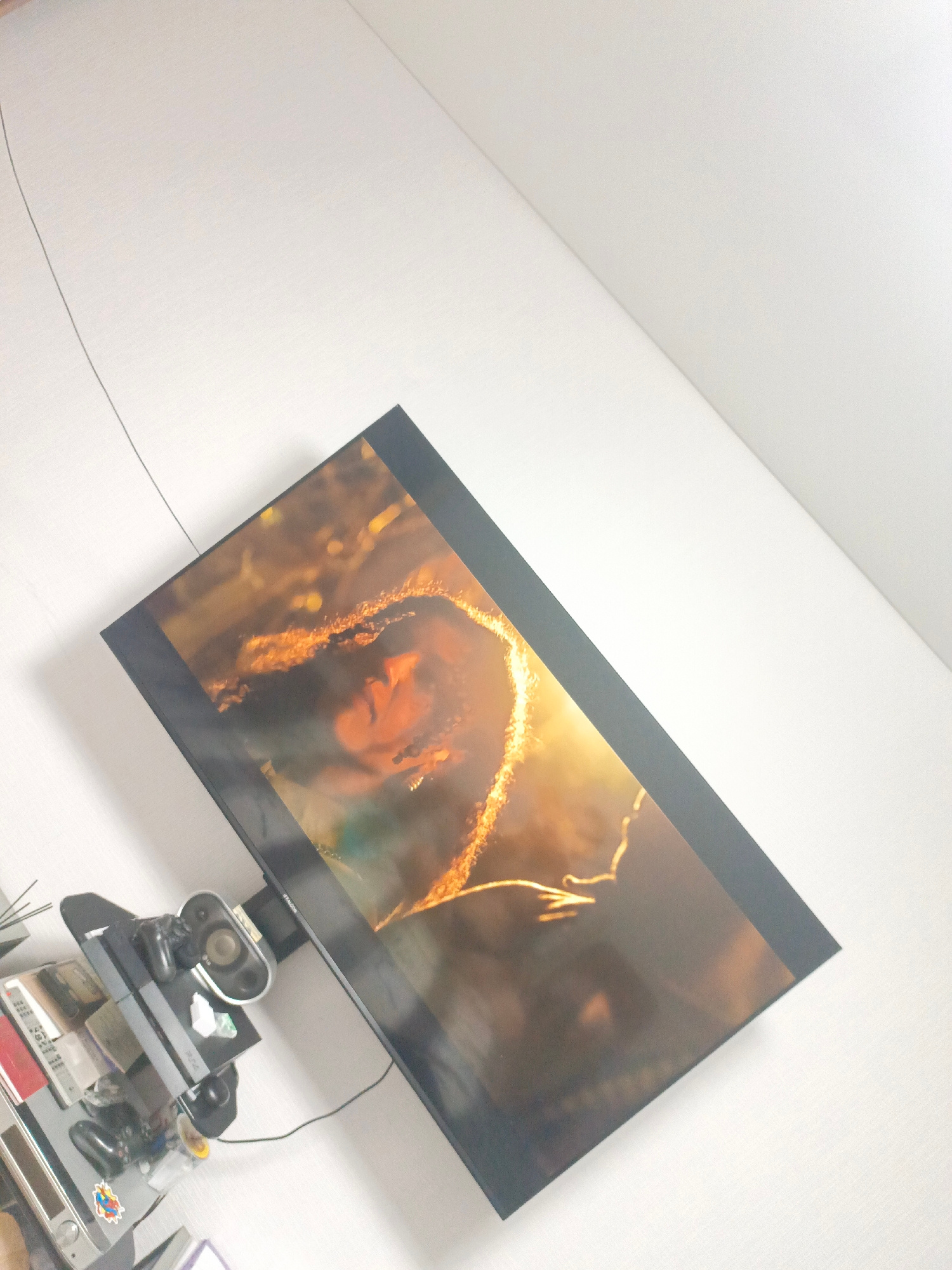 The image shows a large flat-screen television mounted on a white wall, displaying a close-up image of a person with long, dark, curly hair illuminated by a golden light. Below the television, on a black shelf, are various electronic devices, including what appears to be a gaming console with controllers, speakers, and stacks of books or cases. A thin black wire runs from the television up towards the ceiling. The lighting in the room is bright, with light streaming in from the right side, creating a strong contrast and highlighting the texture of the white wall. The overall impression is a dimly lit room with a prominent entertainment setup. There is no information to determine the location in the city of Magadan, Russia.