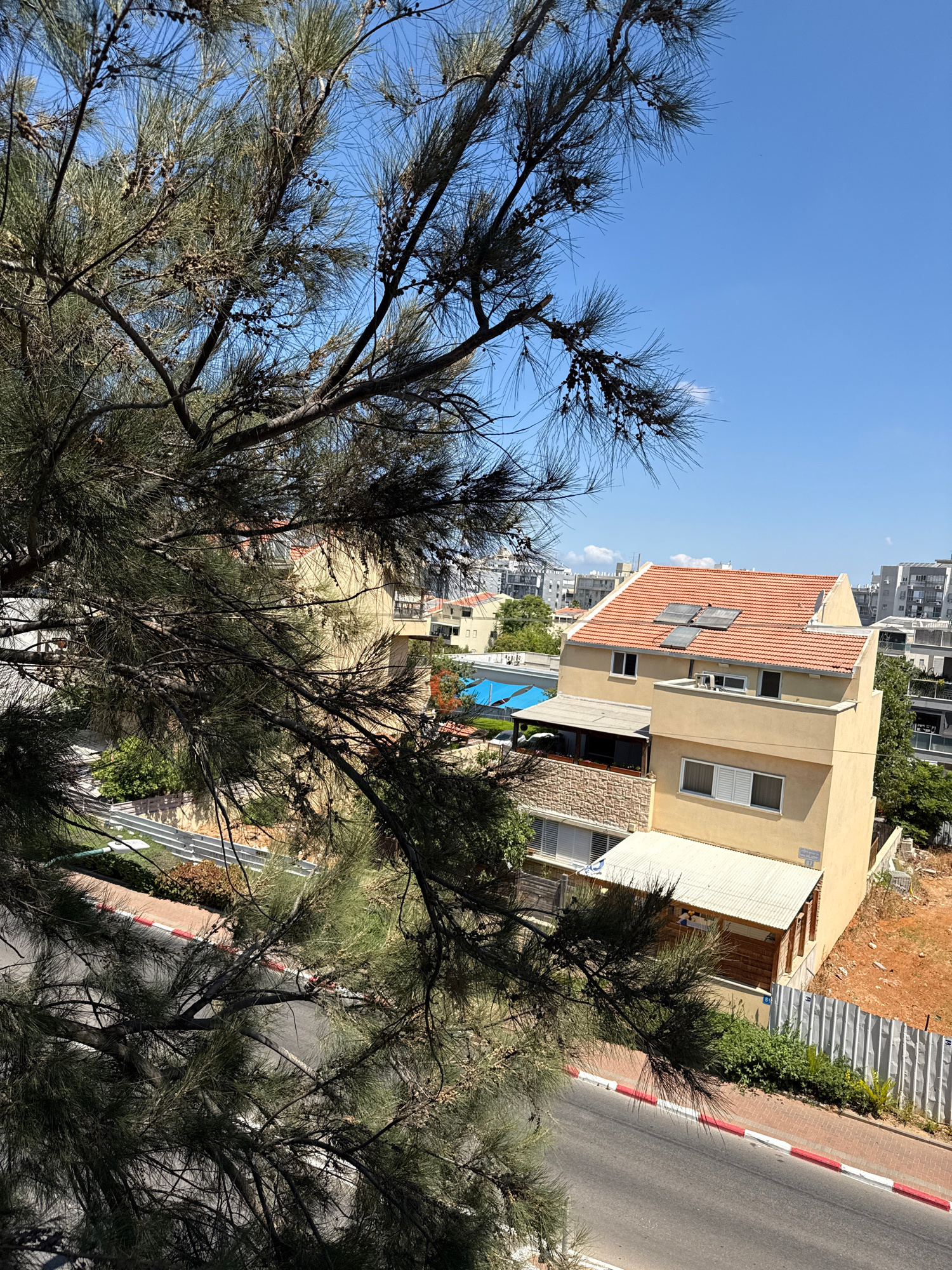 The image, taken from an elevated perspective looking down, captures a scene in Rishon LeZion, Israel, on a clear day. In the foreground, the branches and needles of a pine tree dominate the left side of the frame, partially obscuring the view of the urban landscape behind it.
The midground features a two-story light yellow house with a reddish-orange tiled roof. The roof has several solar panels installed. The house has multiple windows on both floors, with some covered by white shutters. A smaller structure with a corrugated metal roof and wooden siding is attached to the side of the house on the ground level. Adjacent to this structure, a blue swimming pool is visible.
The background displays a typical urban environment with buildings of varying heights and architectural styles. Some of these buildings appear to be residential, while others could be commercial. The sky is a clear, bright blue.
A paved road runs along the bottom of the image, bordered by a sidewalk with red and white markings. A metal fence separates the properties from the road in some areas. The ground on the right side of the image appears to be reddish-brown soil, possibly indicating construction or undeveloped land. Greenery, including trees and bushes, is scattered throughout the scene, adding to the urban-natural mix.