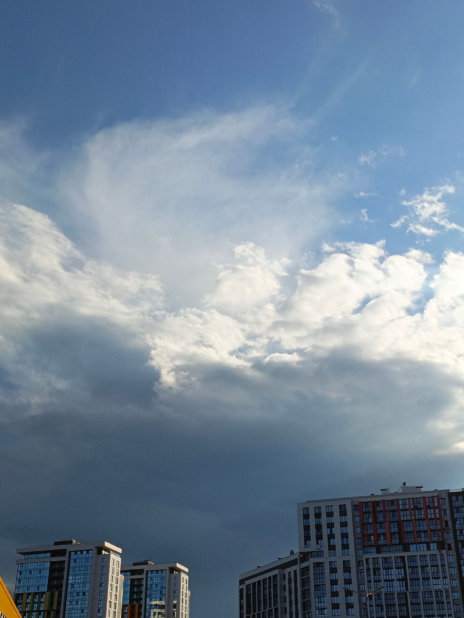 The image captures a dramatic sky over Ufa, Russia, featuring a contrast between bright, white clouds and a darkening, ominous storm cloud formation. The bottom of the frame is occupied by the tops of modern residential buildings, suggesting an urban setting. One building on the right is particularly detailed, with a multi-colored facade featuring orange and white panels interspersed with rows of windows. Another building on the left is lighter in color with blue-tinted windows. The sky is filled with dynamic cloud formations, indicating a change in weather. The light suggests it is either early morning or late afternoon, with sunlight breaking through some of the clouds while others are heavy with rain or storm. No people are visible in the image, and no text is present. The overall mood is one of impending weather change, with a sense of scale provided by the large buildings against the vast sky.