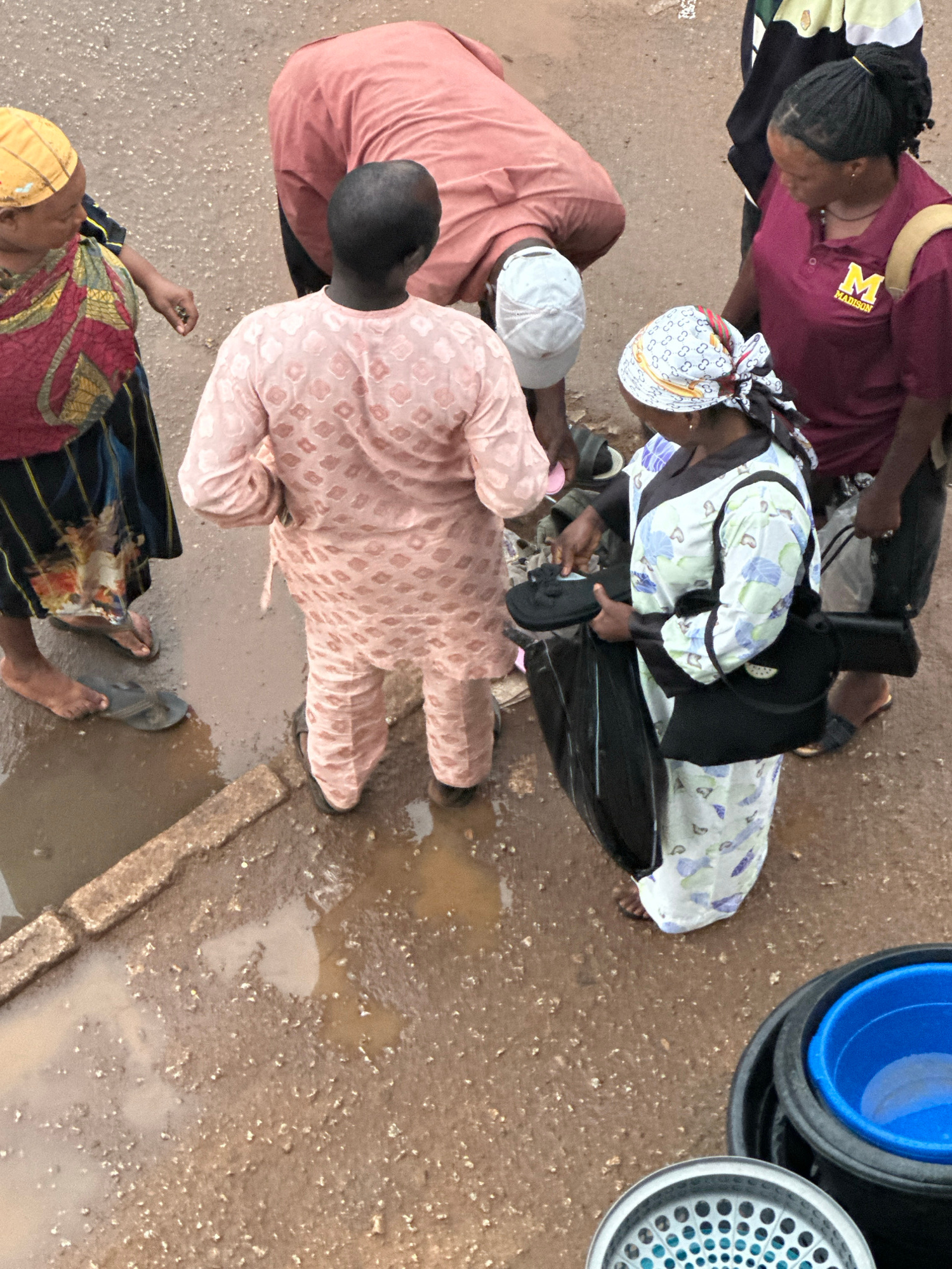 An aerial view shows a group of people gathered on a muddy, uneven surface in Bukuru, Nigeria, likely a street or market area. Several individuals are dressed in traditional Nigerian attire, including a person in a pink kaftan and another in a patterned white and blue outfit. One person wears a maroon shirt with an "M MADISON" logo. The ground is wet and appears to have standing water in puddles, indicating recent rain or poor drainage. Objects visible include black plastic buckets, one containing a blue basin, and a sieve-like metal object. The interaction seems casual, with people engaged in conversation or tending to items.