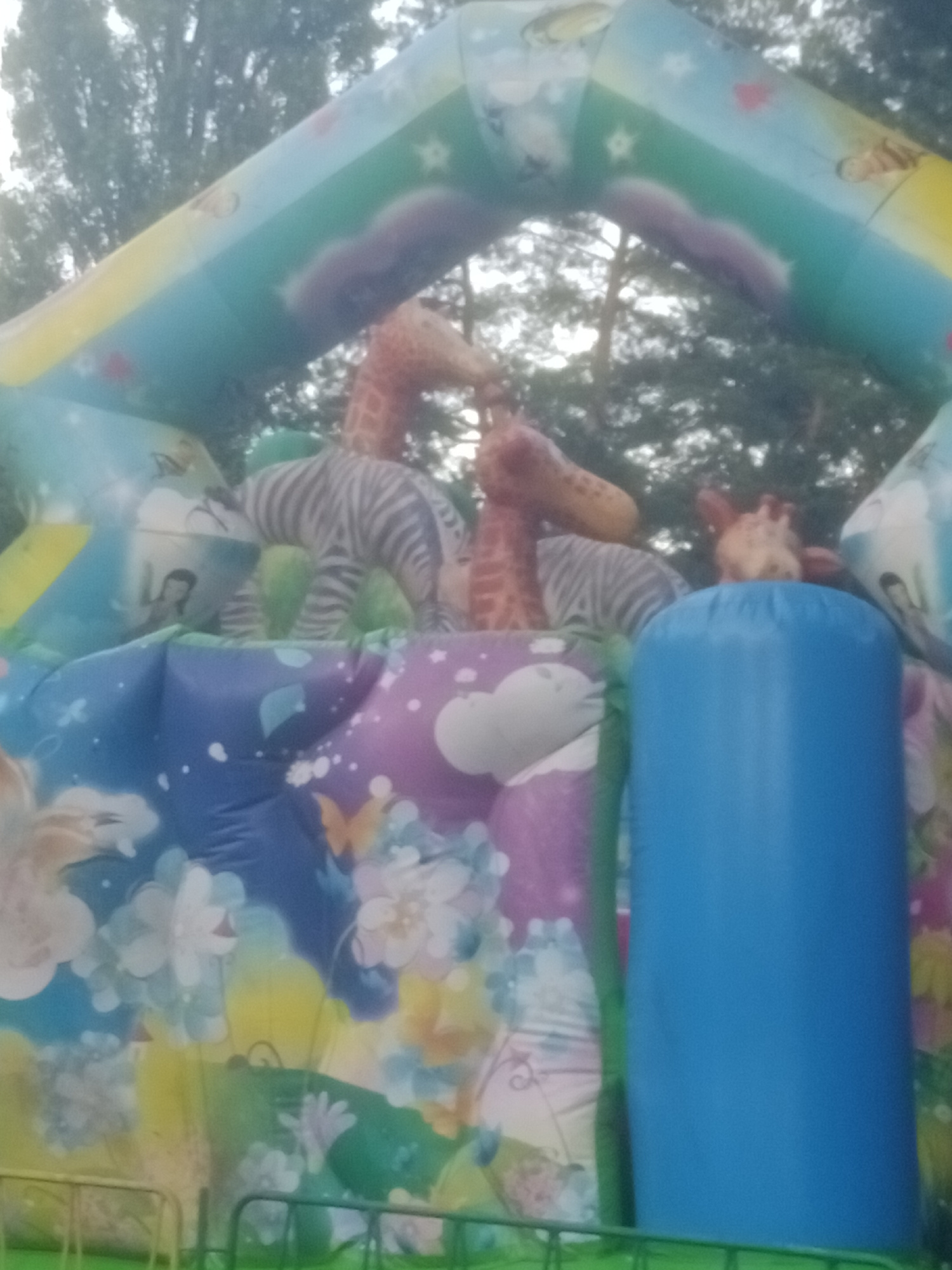 The image captures an inflatable amusement structure, possibly a bounce house or slide, set up outdoors. The structure is adorned with colorful cartoon graphics depicting various animals, including giraffes and a zebra, along with floral patterns and clouds. A large, solid blue cylindrical inflatable is prominently positioned on the right side of the frame. The background shows trees and foliage, suggesting an outdoor location, potentially a park or recreational area within the city of Horishni Plavni, Ukraine. The lighting appears to be natural daylight.