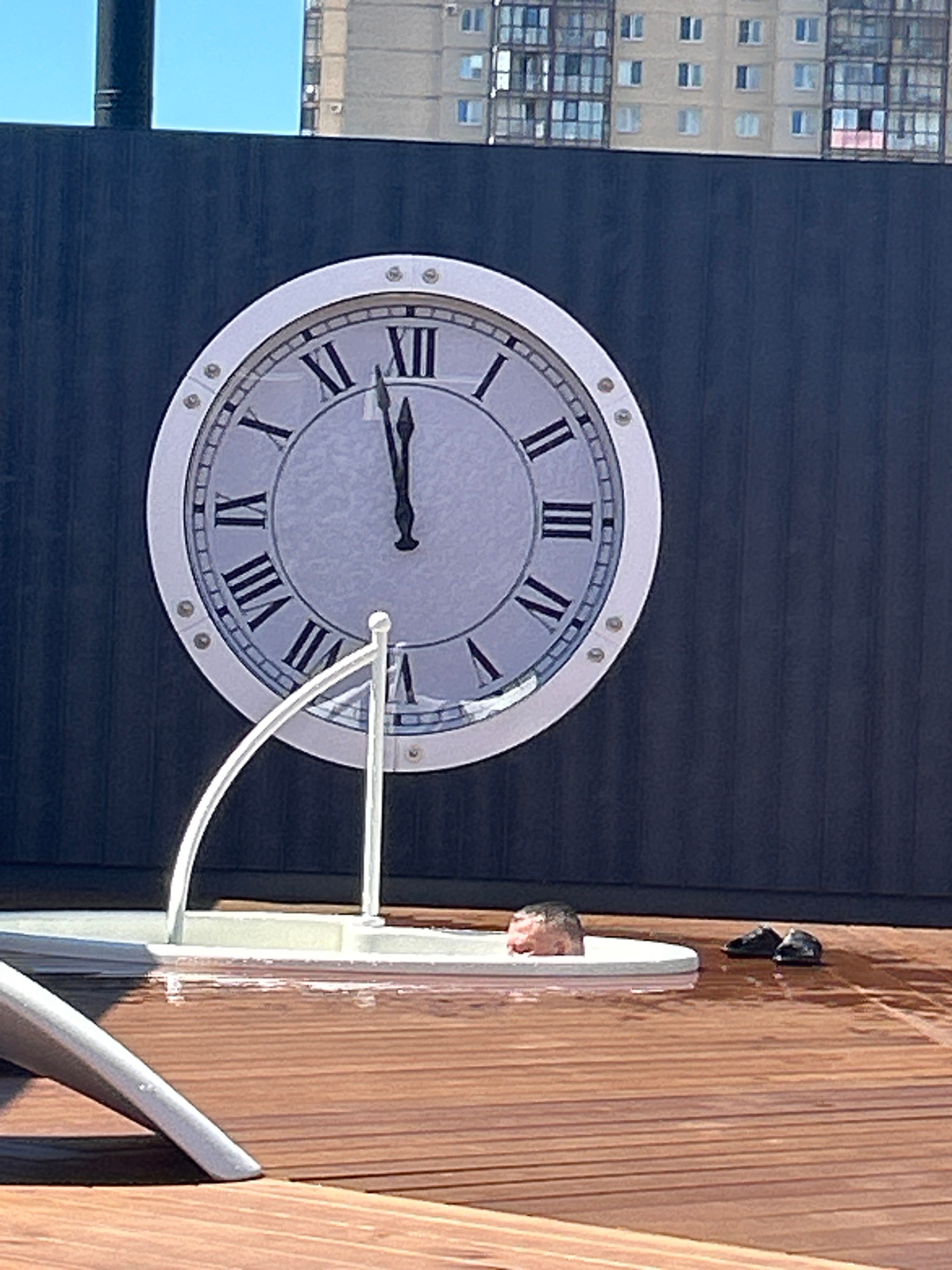 A large analog clock with Roman numerals is mounted on a dark vertical surface. The clock face is light gray with Roman numerals ranging from I to XII. The hour hand is pointing to XII, and the minute hand is pointing to I. Below the clock, a white circular structure is partially filled with water, and what appears to be a pig's head is submerged in the water. To the right of the structure, two black flip-flops are lying on a wet wooden deck. The scene appears to be outdoors, with a building visible in the background under a clear blue sky. The deck is made of wooden planks that are wet, suggesting it may have rained or the area has been recently washed. The overall environment is a contemporary outdoor space, possibly a patio or rooftop.