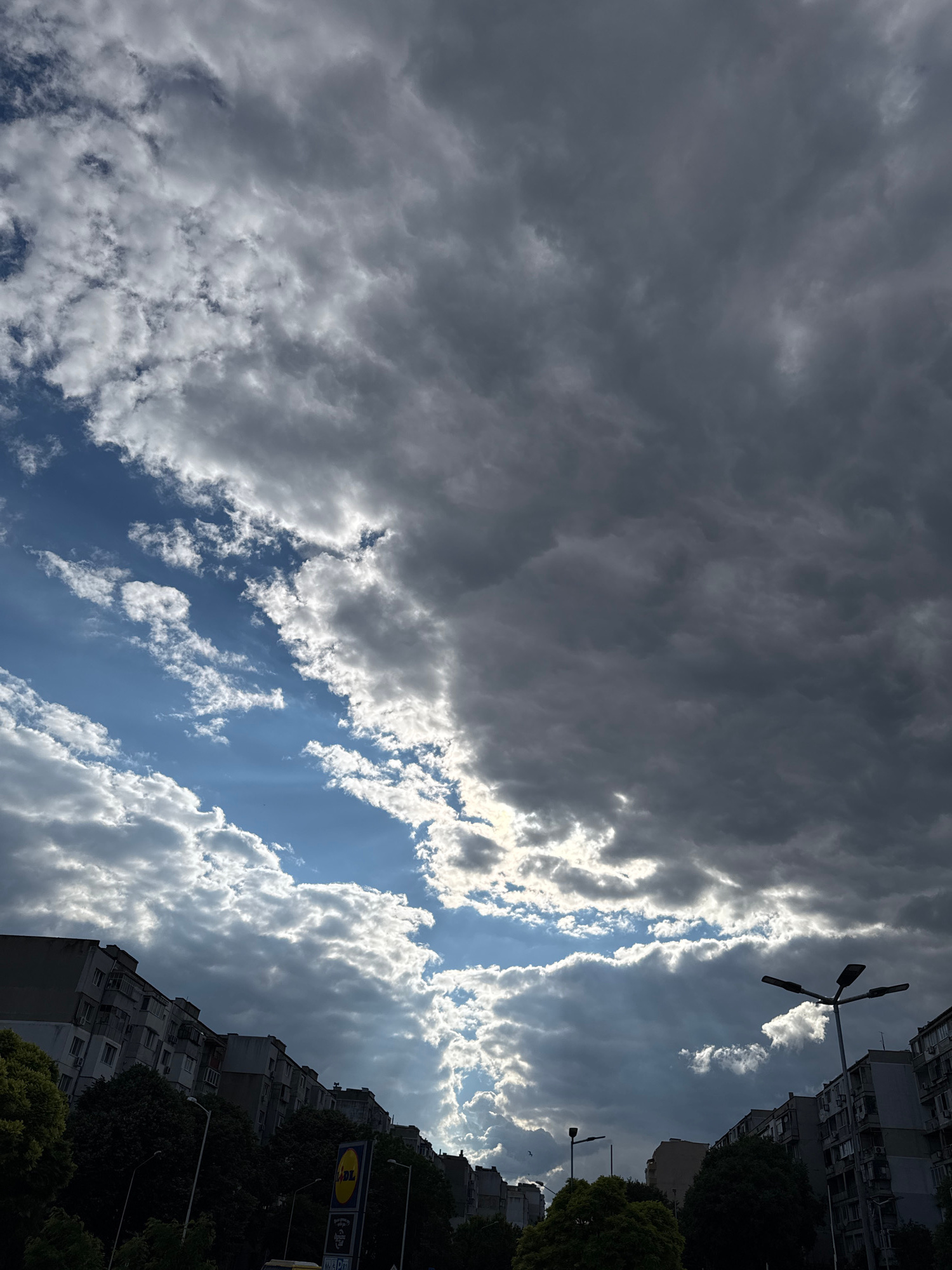 This image depicts a dramatic sky over the city of Varna, Bulgaria. Dark, heavy clouds dominate the upper portion of the frame, interspersed with breaks of bright blue sky and shafts of sunlight piercing through. The lower half of the image shows the rooftops and facades of residential buildings, characteristic of urban Varna. Trees and streetlights are visible along the street level, along with a prominent yellow Lidl supermarket sign. The scene suggests a transition in weather, with a storm potentially approaching or receding.