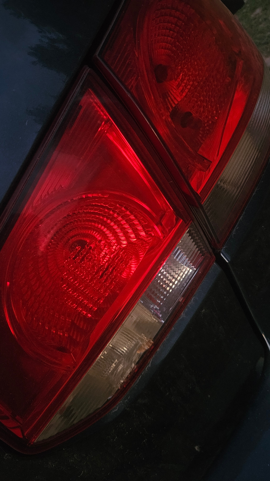 The image shows a close-up, angled view of a car's rear taillight assembly. The taillight is illuminated with a bright red light, revealing a textured, circular pattern within the red lens. Adjacent to the red section, there is a clear or lightly tinted lens with a ribbed or grooved surface. The car's body, which appears to be dark in color, is visible surrounding the taillight assembly. The lighting suggests it is either dusk or night, and the scene is a close-up focus on the automotive component. No location in the city of Unknown, Unknown can be determined from the image.