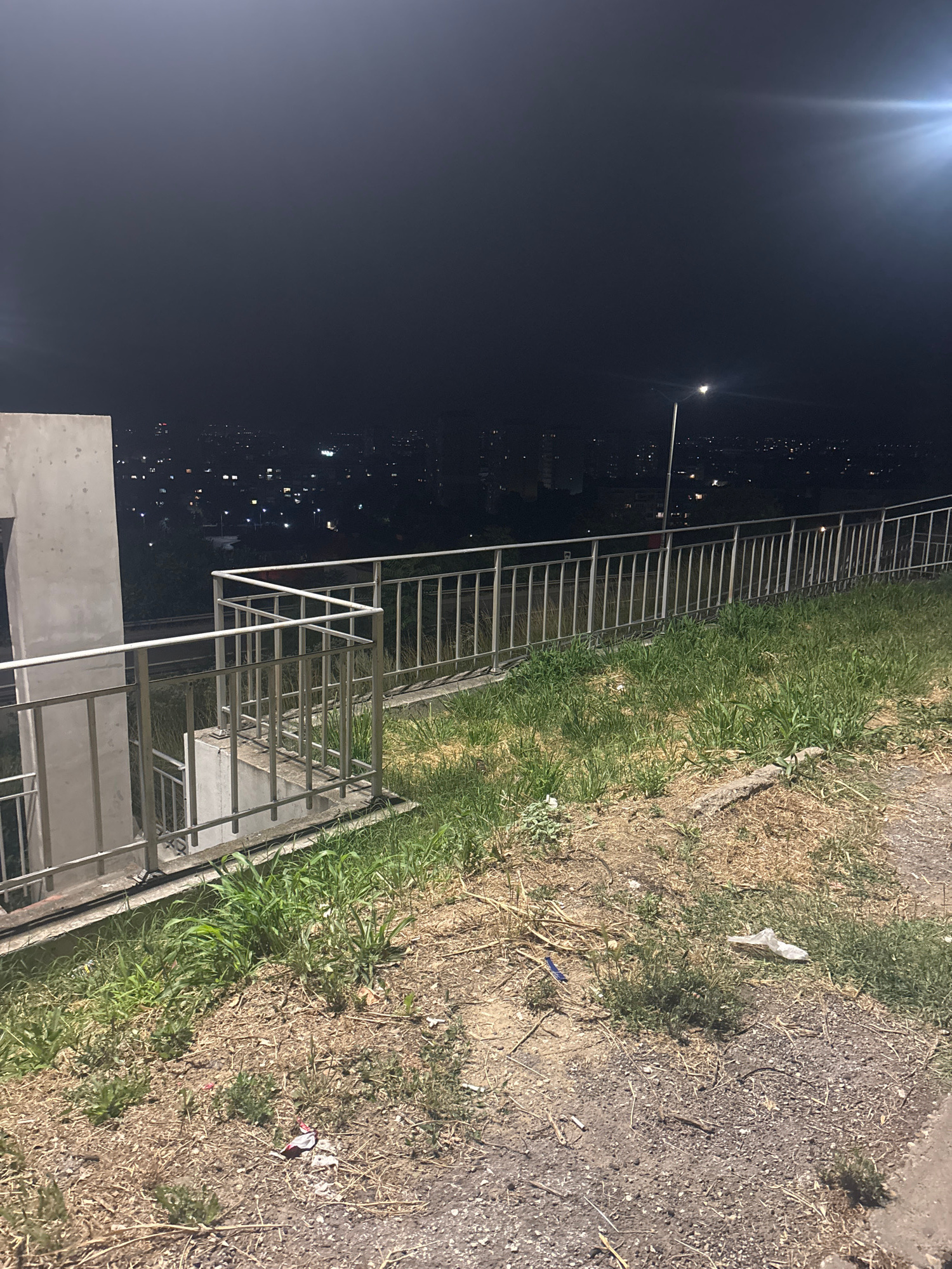 This image depicts a nighttime scene from an elevated vantage point, likely overlooking the city of Ruse, Bulgaria. In the foreground, a concrete structure and a metal railing line a grassy embankment. The railing appears to be part of a walkway or viewing platform. Beyond the railing, the distant city lights are visible, suggesting a populated area. A streetlamp is illuminated in the mid-ground, casting a bright light. The sky is dark, with a lens flare effect visible in the upper right quadrant, possibly from a bright light source such as the moon or a strong artificial light. The ground in the foreground is covered with dry grass and some scattered debris, including what appears to be litter.