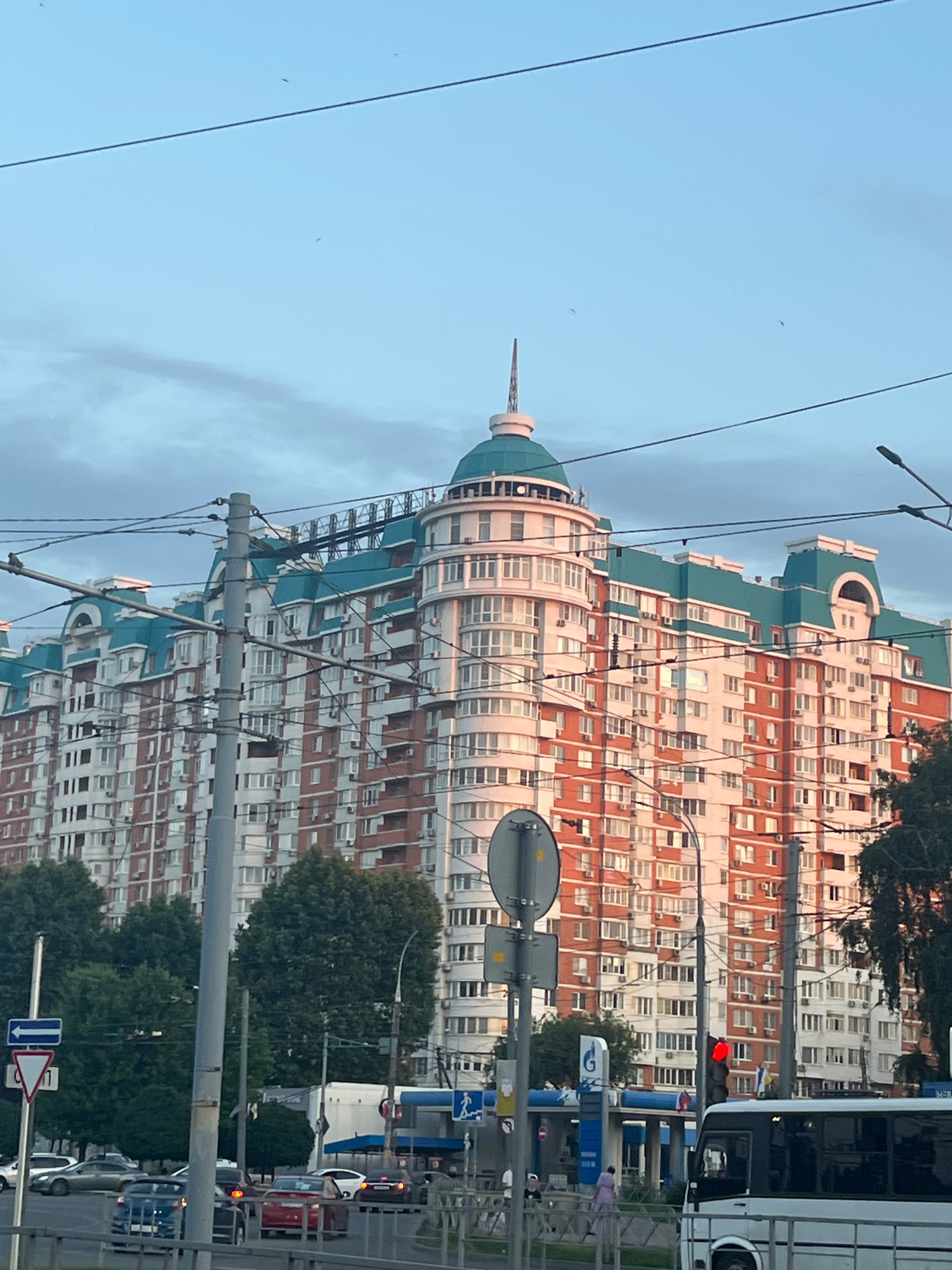 The image captures a large, multi-story residential building with a distinctive turquoise roof and red-brick facade. The building features numerous windows and balconies. A rounded, turret-like structure topped with a spire is prominent on the left side of the building's facade. Overhead, electrical wires and utility poles crisscross the sky, with a few birds visible in the distance. In the foreground, a street scene unfolds with several cars, a bus, traffic lights, and directional signs. Lush green trees partially obscure the lower levels of the building on the left. The lighting suggests it is either sunrise or sunset, casting a warm glow on the building. This building is identified as the "Malakhit" residential complex located at 30-36 V. E. Klyucheva Street in Krasnodar, Russia.