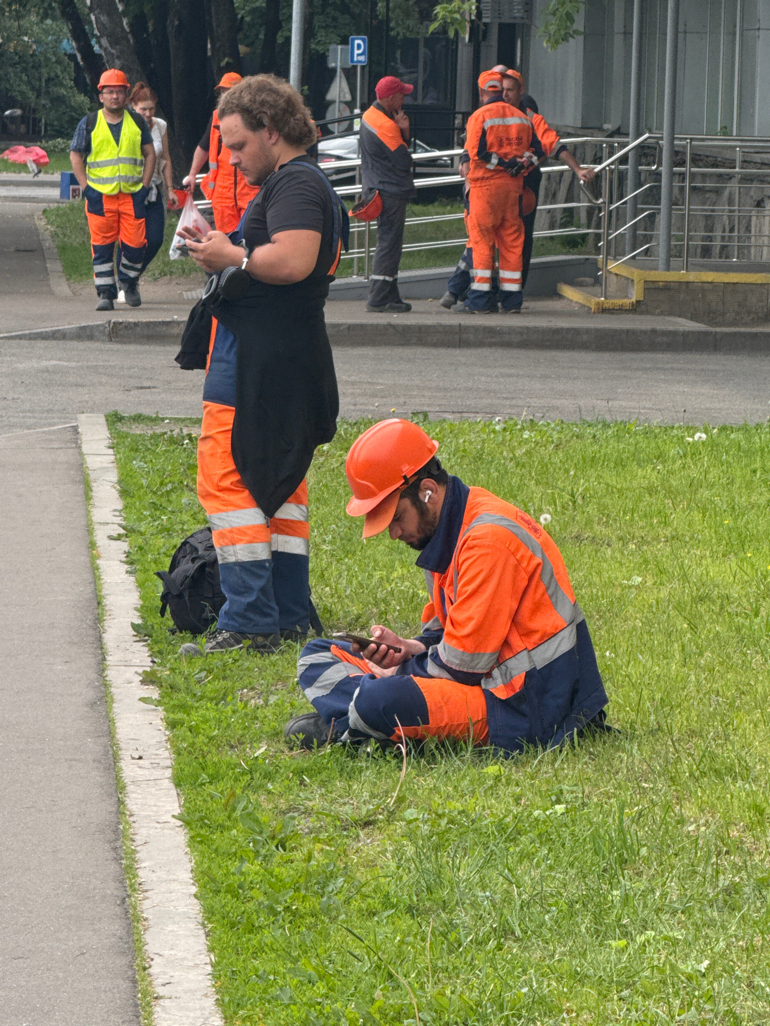 In Moscow, Russia, a group of workers in bright orange and blue uniforms are visible. Two men are standing and looking at their phones. One worker, wearing a yellow safety vest over his uniform, stands with his hands in his pockets, while another worker stands nearby. In the foreground, a man in an orange hard hat and uniform is sitting on the grass, looking at his phone. Another worker is also visible in the background, talking to a colleague. The environment appears to be a street or sidewalk next to a grassy area, with some trees and a building in the background. The overall scene depicts a moment of pause and rest for the construction workers.