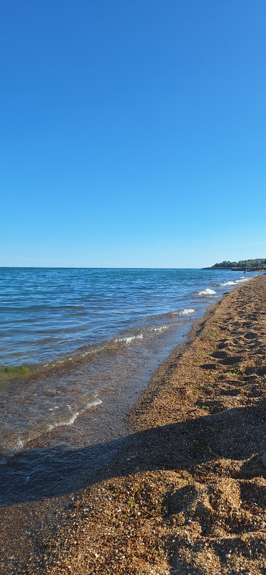 A wide-angle, eye-level vertical shot captures a serene beach scene under a clear, bright blue sky. The ocean, a deep blue, stretches to the horizon, meeting the sky in a sharp, defined line. Gentle waves with white crests lap at the shore, creating a shimmering effect on the water's surface. The beach itself is composed of fine, light brown sand mixed with small pebbles and some sparse green vegetation. A distinct shadow stretches across the foreground, cast by an unseen object. In the distance, a hilly coastline with trees is visible on the right side of the frame. There are no people or animals present in the image.