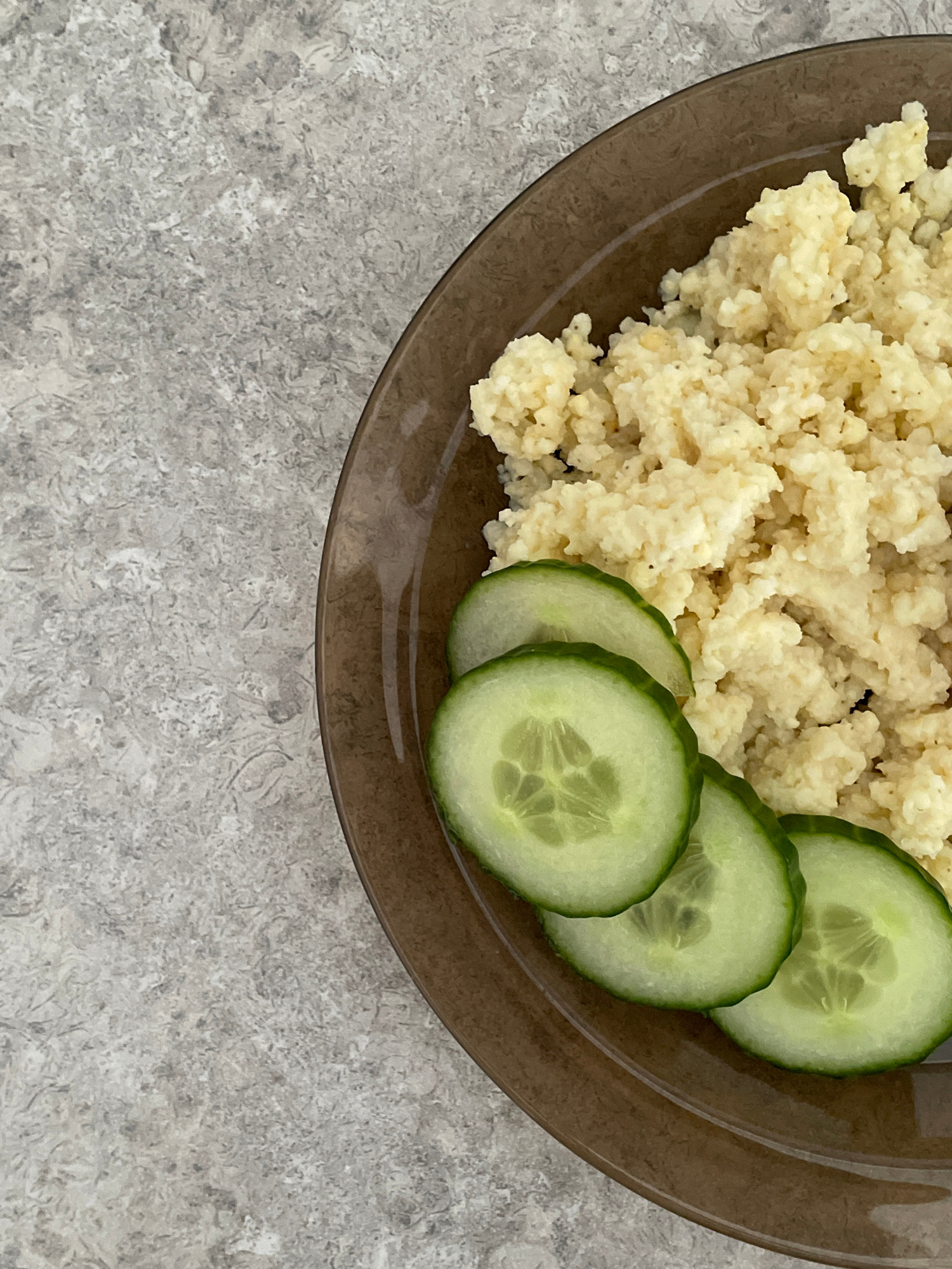 A close-up, overhead shot captures a brown, translucent plate filled with a mound of couscous and three slices of cucumber. The couscous is light yellow and has a fluffy, granular texture. The cucumber slices are green with visible seeds and are arranged in a slight overlap to the left of the couscous. The plate rests on a speckled, light gray surface that resembles a stone countertop or placemat. The lighting is even, with no strong shadows, and the focus is sharp on the food and plate. The background is blurred and out of focus.