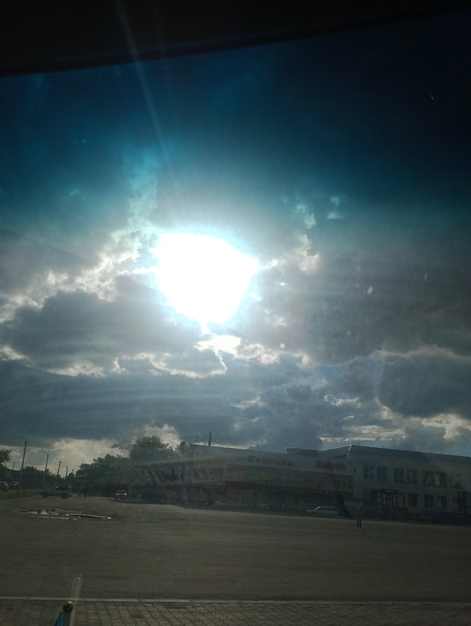 The image captures a cloudy sky with the sun shining brightly through a gap in the clouds, casting rays of light downwards. Below the sky, a street scene is visible, featuring a building with multiple storefronts, including "Sinsay" and "Shikpak." Several cars are parked or driving on the street and in front of the building. Trees line the street in the background. The foreground shows a paved area, possibly a parking lot or a pedestrian walkway, with some visible puddles of water. The overall impression is of an outdoor commercial area in Atbasar, Kazakhstan, under a dramatic sky.