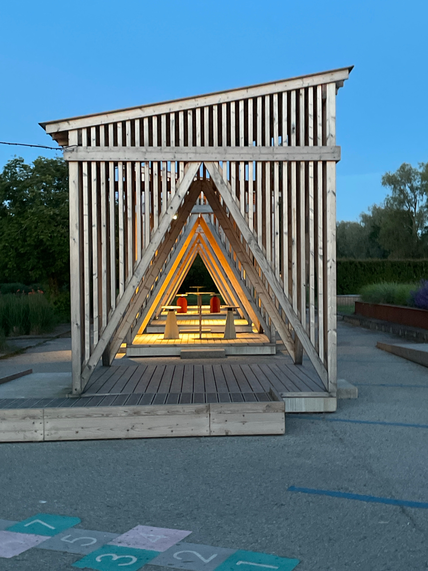 An open-air wooden structure with a triangular prism shape is illuminated from within by warm lights. The structure is made of vertical wooden slats forming the sides, with diagonal wooden beams supporting the roof. Inside, there are two red seating elements and two small tables. The structure is situated on a wooden deck, which is part of a larger paved area. In the foreground, there are chalk markings on the pavement resembling a hopscotch grid with numbers. The background shows greenery, including trees and bushes, and a clear blue sky. The lighting suggests it is either dawn or dusk. The scene appears to be in a public outdoor space, possibly a park or recreational area, located in Tallinn, Estonia.