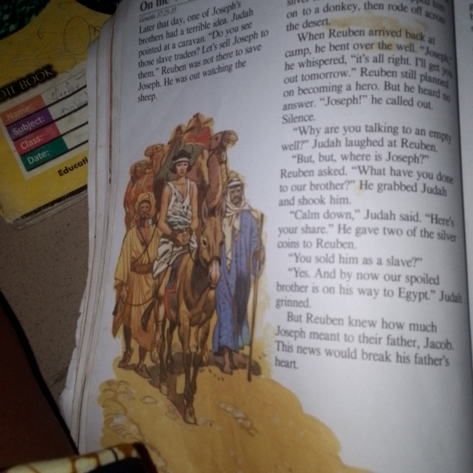 The image shows an open book with text and an illustration. The text is from the Bible, specifically Genesis 37:25-35, and depicts a story about Joseph and his brothers. The illustration shows a group of men and camels in a desert landscape. Some of the men are riding camels, while others are walking. The men are dressed in traditional Middle Eastern clothing. The text on the left side of the book is a label with fields for "Name," "Subject," "Class," and "Date." This label appears to be from a notebook or a school-related item. The overall scene suggests a religious or educational context. There is no indication of a specific city or location.