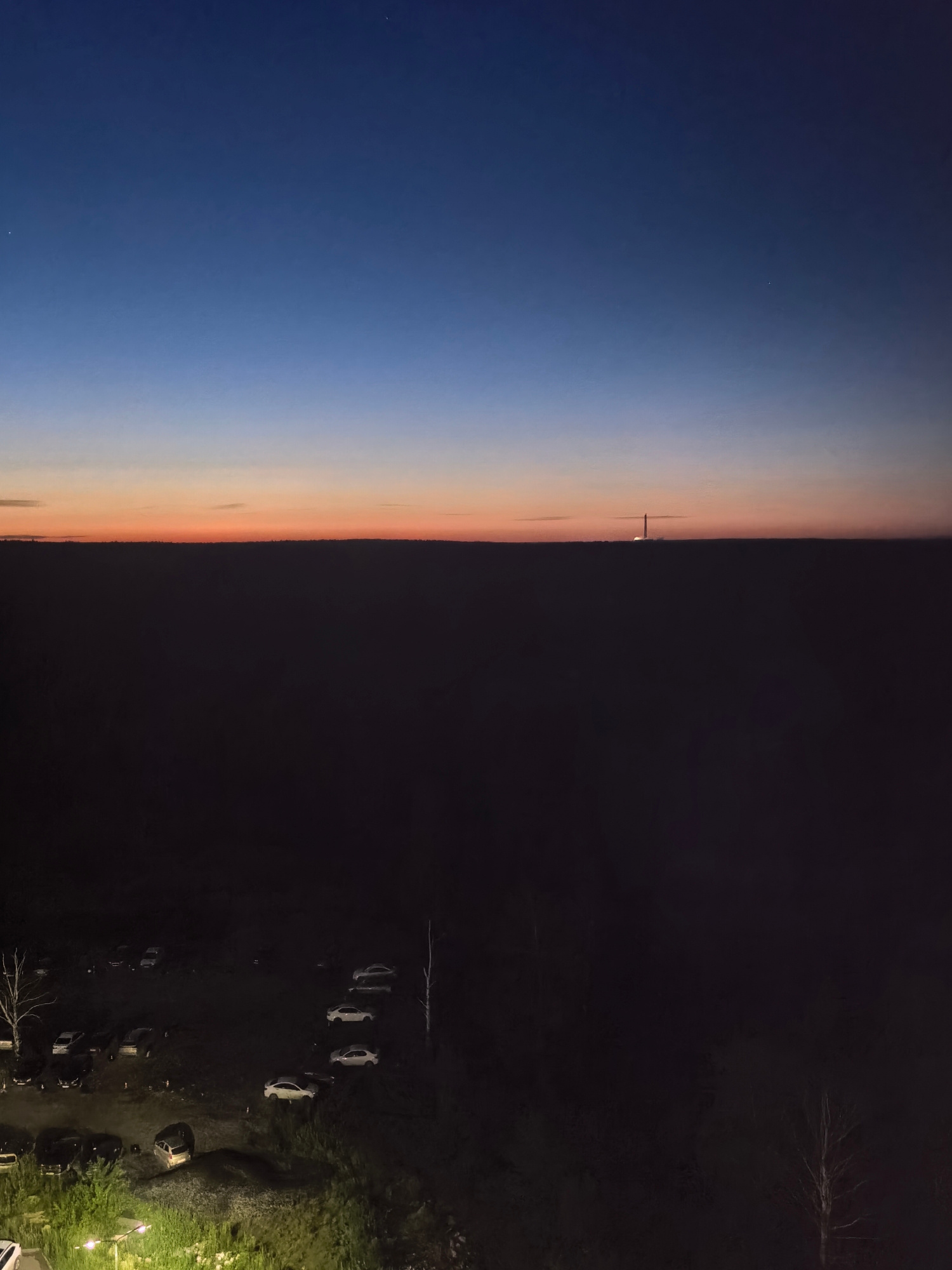 A wide-angle, high-angle shot captures a dim, dusky landscape with a vibrant gradient of sky fading from deep blue to a thin band of orange along the horizon. A dark, silhouetted landmass stretches horizontally across the frame, obscuring most of the ground. A faint, vertical structure, possibly a tower or antenna, is visible in the distance on the horizon. In the lower portion of the frame, a dimly lit parking lot is visible with several cars parked. The ground is covered in dark vegetation, with some bare trees scattered throughout the scene. A single streetlamp illuminates a section of the foreground, casting a greenish glow on the surrounding grass and some parked vehicles. The overall impression is one of twilight, with the last vestiges of daylight giving way to the darkness of night.