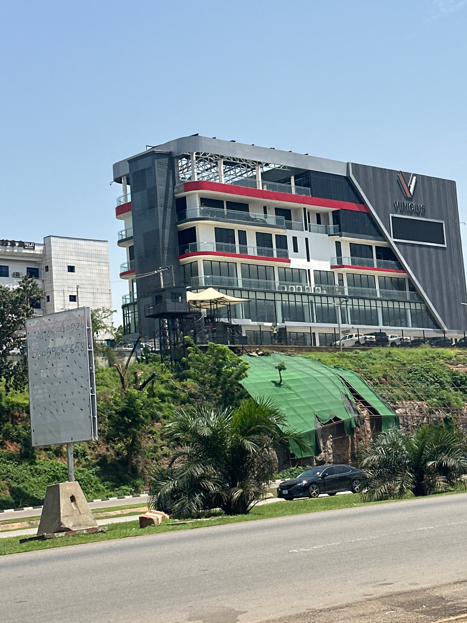 The image shows a modern, multi-story building with a distinctive architectural design, featuring a dark gray facade with angled lines and red accents on the upper floors. The building has a large "V VINCIUS" logo on its upper right side. Below this building, a green tarp covers a section of a hillside, appearing to be part of a construction or repair site. In the foreground, a paved road runs horizontally across the bottom of the frame, with a grassy embankment and some vegetation between the road and the building. A dark sedan is parked on the grassy area next to the road. A large, light-colored billboard with some handwritten numbers is also visible on the left side of the image. In the background, another building, lighter in color and appearing older, can be seen. The sky is clear and blue, indicating a sunny day. The scene is likely located in a commercial or urban area of Abuja, Nigeria, given the presence of modern architecture and signage consistent with the city.