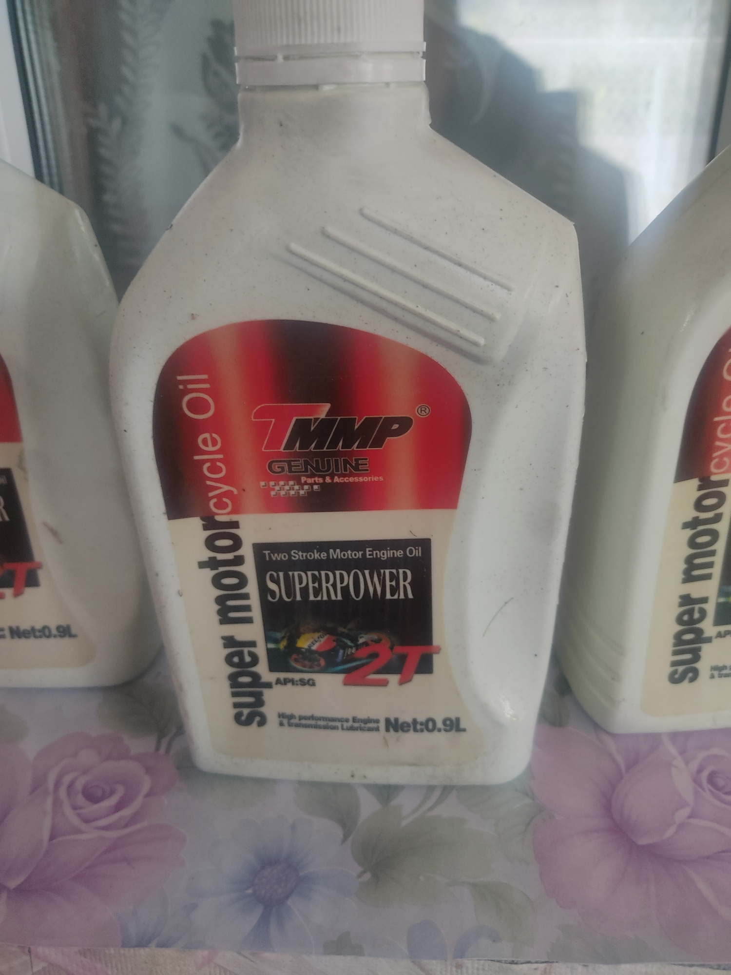 This media file features two white plastic bottles of motorcycle engine oil, stacked one on top of the other. The bottles are positioned at an angle, with the top bottle partially obscuring the bottom one. Both bottles have labels that are largely identical. The prominent text on the labels reads "super motorcycle Oil" and "SUPERPOWER". There is also a logo that appears to be "TMP" and "GENUINE Parts & Accessories". The labels also indicate "Two Stroke Motor Engine Oil", "API:SG", and "Net:0.9L". The background of the image is a surface with a floral pattern, featuring shades of pink, purple, and green, resembling a tablecloth or decorative paper. The overall lighting suggests an indoor setting, possibly a shelf or table. The image is a still photograph, and no specific activity or event is depicted beyond the presentation of the products. The condition of the bottles suggests they are new or unused, as there are no visible signs of wear or damage. There are no people or animals visible in the image.