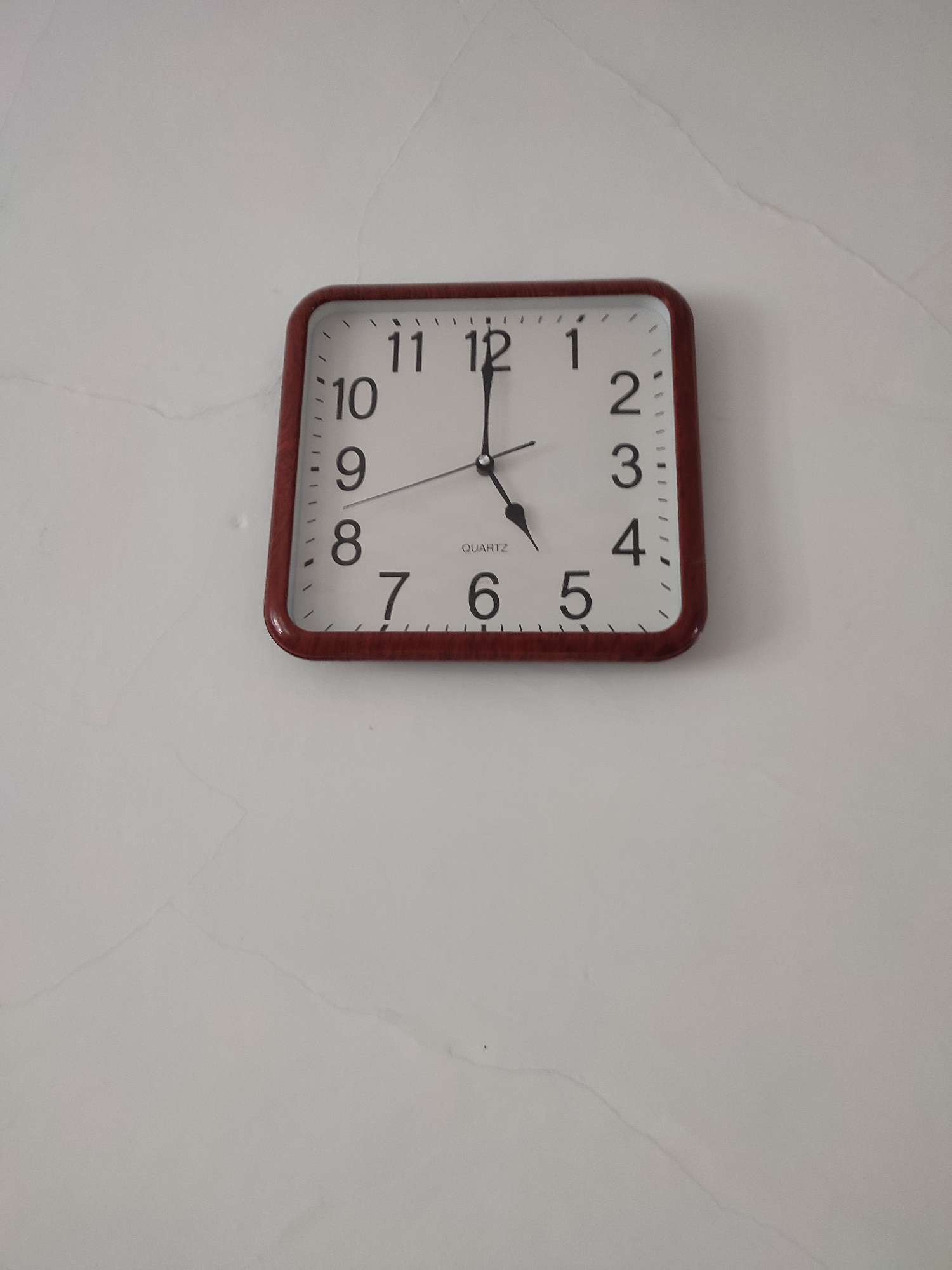 A square clock with a dark brown frame hangs on a white, slightly textured wall. The clock face is white with black Arabic numerals indicating the hours. The minute hand points to the 12, and the hour hand points to the 5. The wall appears to be a neutral color and has some faint lines or cracks visible. The overall scene is indoors. The location in Altai, Kazakhstan is not discernable from the image.