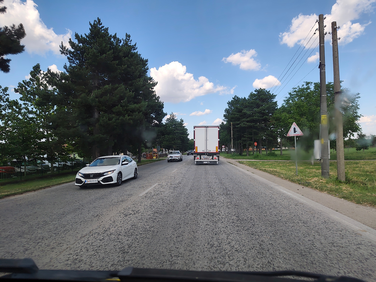 The image captures a forward-facing view from a vehicle driving on a paved road, likely within or near Obnova, Bulgaria. The road is bordered by a grassy verge and lined with trees on both sides. In the foreground, the dashboard of the vehicle is partially visible.
Several vehicles are present on the road. A white sedan, possibly a Honda Civic, is in the left lane, closer to the foreground. Further ahead, a white truck with a red stripe is in the right lane. Another white car, a hatchback, is visible behind the sedan, and a darker vehicle is behind the truck.
The sky is a vibrant blue with scattered white clouds. Utility poles with power lines are visible along the right side of the road. A triangular road sign with a black symbol depicting a road splitting is also present near the right side of the road. The overall impression is a daytime scene on a relatively clear day, with moderate traffic.