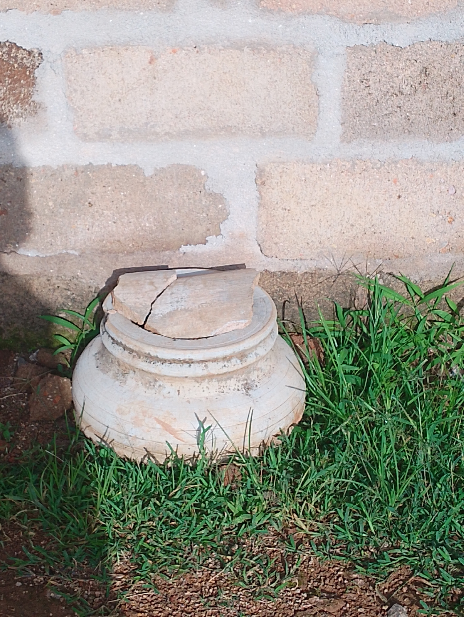 The image shows a broken, light-colored ceramic pot lying on the ground next to a brick wall. The pot appears to be a traditional African design with a rounded body and a flared rim, although much of the upper section is missing. Two large pieces of the broken rim are resting on top of the pot, one of which is cracked. The pot is surrounded by green grass and a patch of reddish-brown soil. Behind the pot, a section of a brick wall is visible. The bricks are rectangular and light brown in color, with lighter gray mortar between them. The wall appears to be weathered, with some patches of missing mortar and a stained appearance. The lighting suggests it is daytime, with shadows cast on the wall and grass. The overall scene evokes a sense of abandonment or neglect, with the broken pot and overgrown grass indicating a lack of recent activity. No people are visible in the image, and there is no visible text. The context of Russo, Nigeria, suggests a warm, possibly tropical climate.