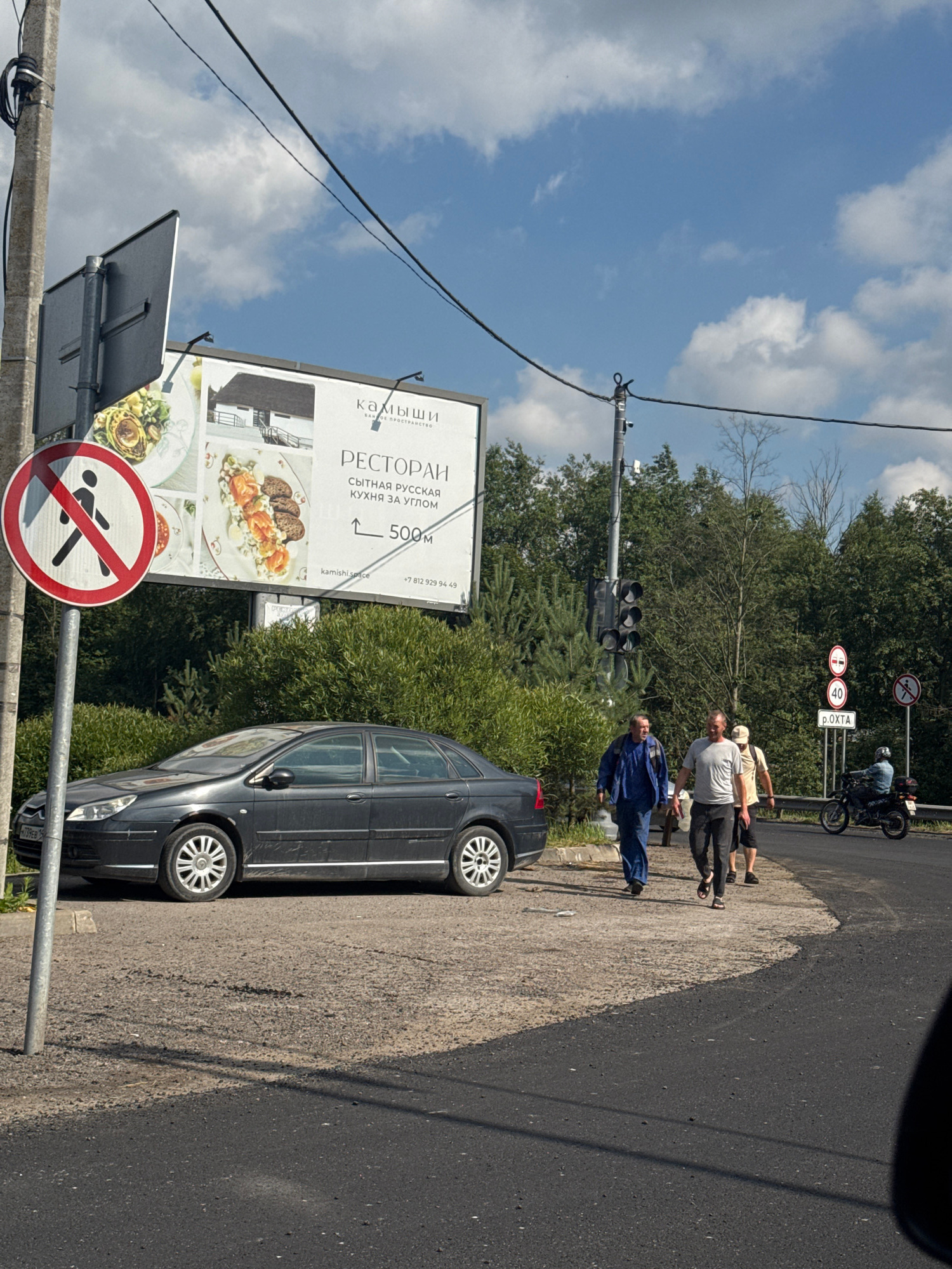The image depicts a roadside scene on a sunny day, likely in or near Городок, Russia. A dark-colored sedan is parked on the side of a paved road, next to a gravel shoulder. In the background, a dense line of green trees forms a natural border. A large billboard advertises a restaurant named "Камыши" (Kamyshi), featuring images of food and offering "Russian cuisine 500m away." A traffic light is visible to the left of the billboard.
Several people are present. Two men are walking away from the camera on the side of the road. One is wearing a blue jumpsuit, and the other is in a light-colored t-shirt and dark pants. Further down the road, a person on a motorcycle is visible.
A prominent "no pedestrians" sign, a red circle with a diagonal line across a pedestrian silhouette, is mounted on a pole to the left of the billboard. Other road signs are also visible, including a speed limit sign of "40" and a sign indicating "p. OKHTA" (presumably a settlement or location). The sky is blue with scattered white clouds. The overall environment suggests a rural or semi-rural roadside area.