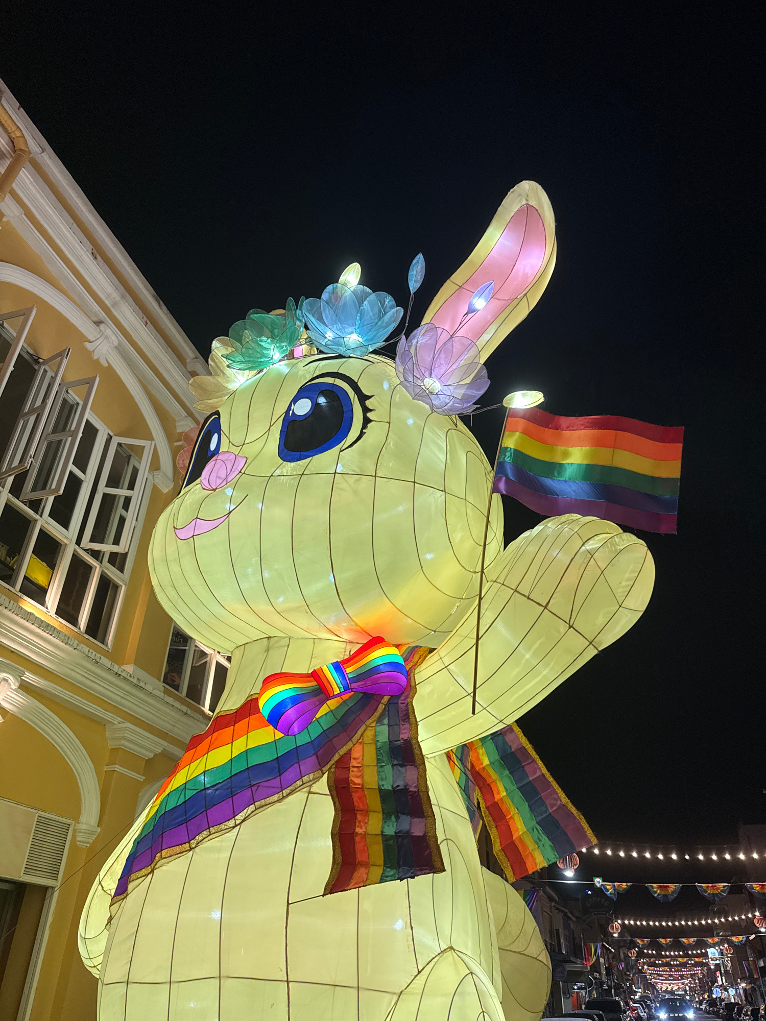 A large, illuminated rabbit lantern, adorned with rainbow-colored accessories and a rainbow flag, stands prominently at night in Phuket City Municipality, Thailand. The lantern is a pale yellow and features large blue eyes, a pink nose, and a cheerful smile. It is decorated with lotus-shaped lights on its head and a rainbow bow tie and scarf around its neck. In its right paw, it holds a rainbow flag. The lantern is positioned in front of a historic yellow building with open windows and white architectural details. In the background, a street is visible, lined with buildings and illuminated by strings of festive lights. Cars are parked along the street and traffic lights are visible. The scene suggests a festive event or celebration in Phuket, likely related to LGBTQ+ pride.