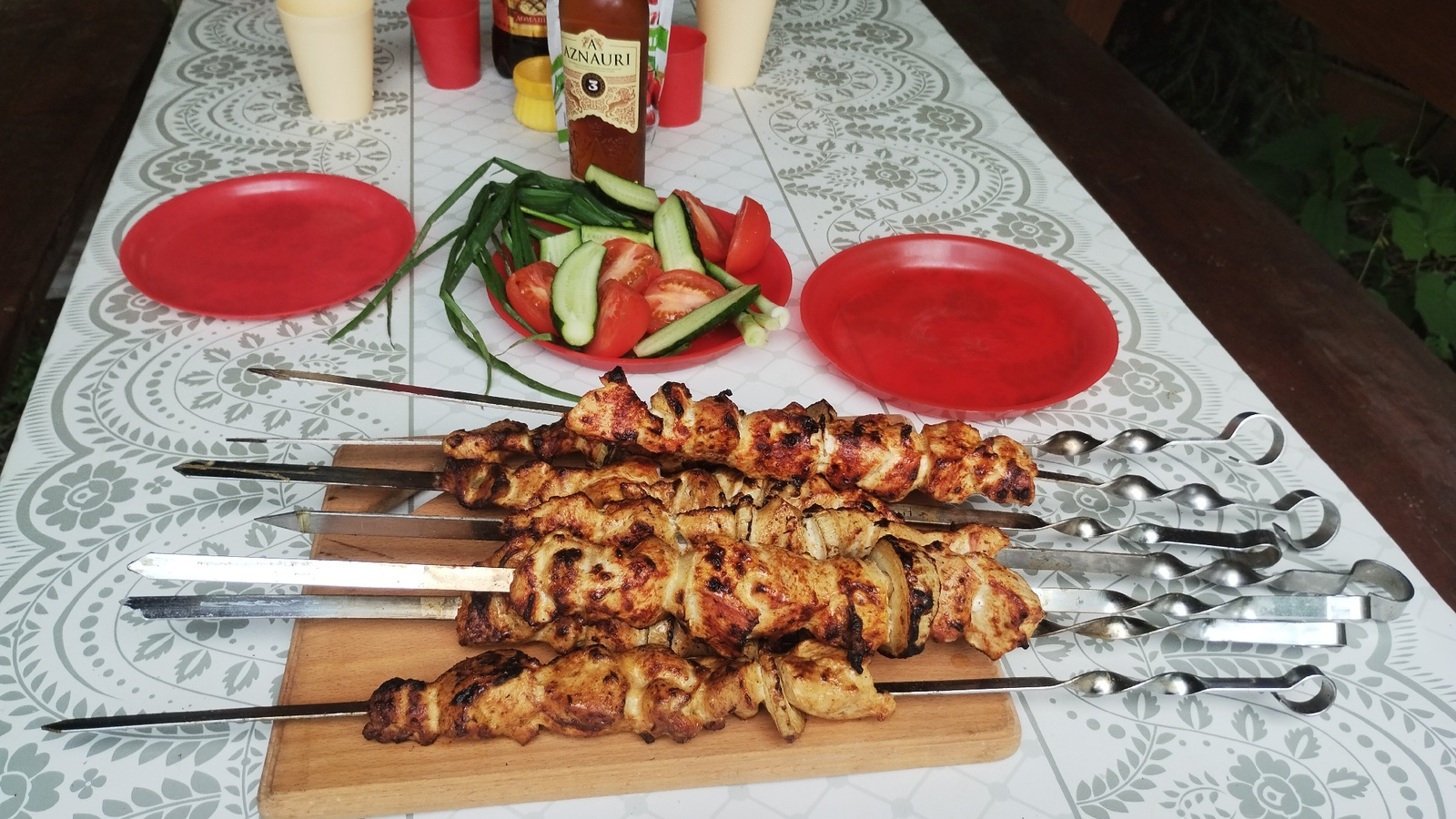 The image depicts a table laden with food, set outdoors on a patterned tablecloth. In the foreground, four skewers of grilled chicken shashlik rest on a wooden board. To the left and right of the shashlik are two red plates, one empty and one containing a salad of sliced tomatoes and cucumbers with green onions. In the background, several cups, a bottle of Aznauri brandy, and other items are visible. The setting appears to be a picnic or outdoor meal in Synevyrska Poliana, Ukraine.