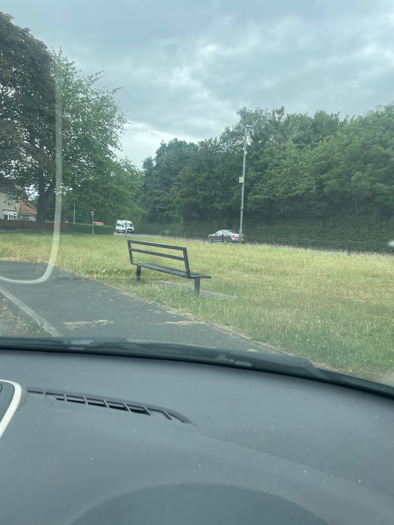 The image shows a view from inside a vehicle, looking out at a park-like area with a bench. The foreground is dominated by the dashboard of the car, including a vent. Beyond the vehicle, there is a path, a grassy area with a bench, and trees. A dark-colored car is parked or driving on a road in the distance, and a white van is also visible. The sky is overcast.