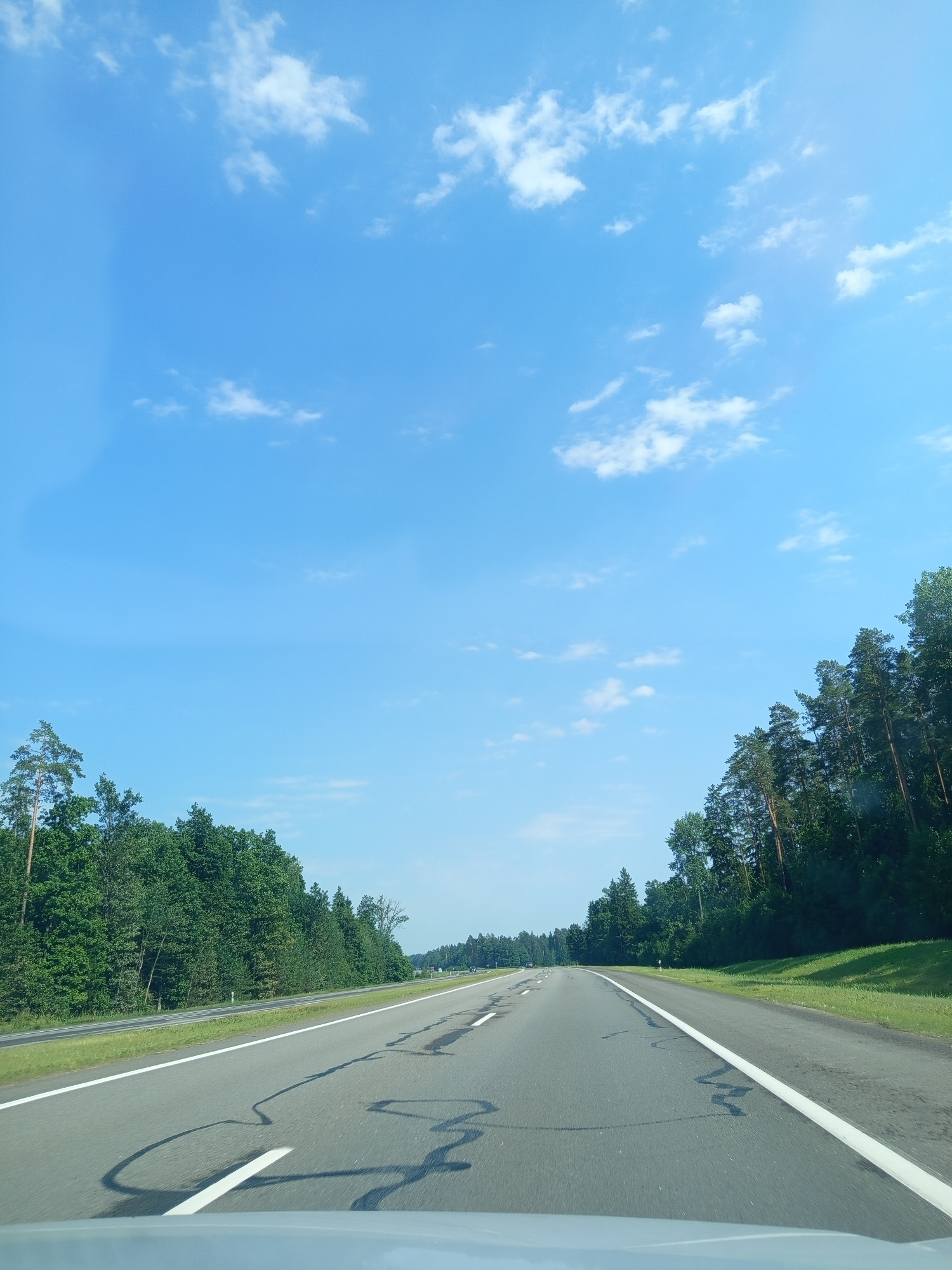This media file captures a view from a moving vehicle on a bright, sunny day. The scene is dominated by a clear blue sky with scattered white clouds, suggesting fair weather. To the right of the frame, a paved road stretches into the distance, bordered by a white line. The road surface appears to have some dark, irregular markings. Adjacent to the road, on the left side of the image, is a dense line of lush green trees, primarily conifers, forming a forest. Beyond the trees, the landscape slopes gently upwards, covered in grass. The perspective indicates the photo was taken from inside a vehicle, possibly from the passenger seat or dashboard, looking forward and slightly upwards. The orientation of the image is rotated, making the sky appear on the left and the road on the right. No people or specific objects other than the natural environment and the road are visible. The time of day appears to be daytime, given the bright sunlight and clear sky. The location context provided is Valeŭačski Sieĺski Saviet, Belarus.