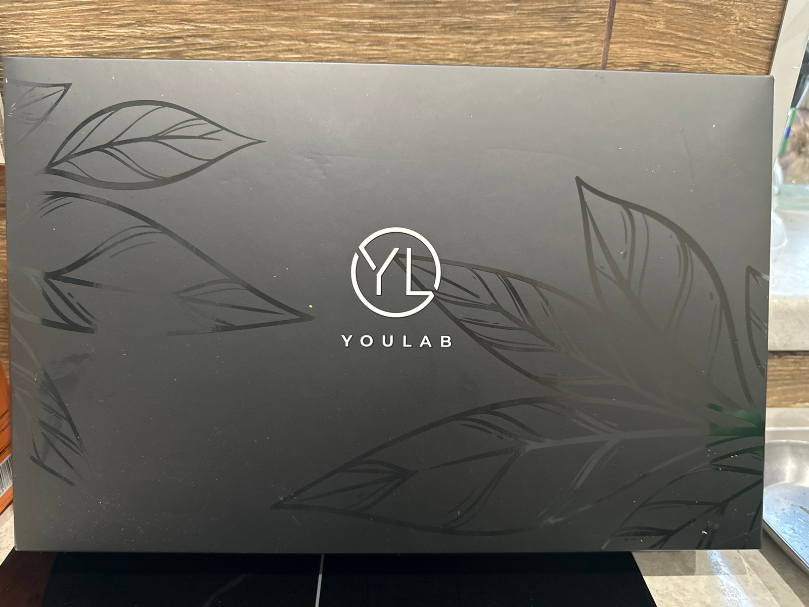 The image shows a closed, dark gray rectangular box with a minimalist design featuring outlines of leaves and the embossed text "YOULAB" with a circular logo above it. The box appears to be on a dark surface, possibly a table, with a wooden-textured background visible behind it. There are some specks of dust or debris on the surface of the box. The lighting suggests an indoor setting. There is no information in the image that indicates the location in the city of Ufa, Russia.