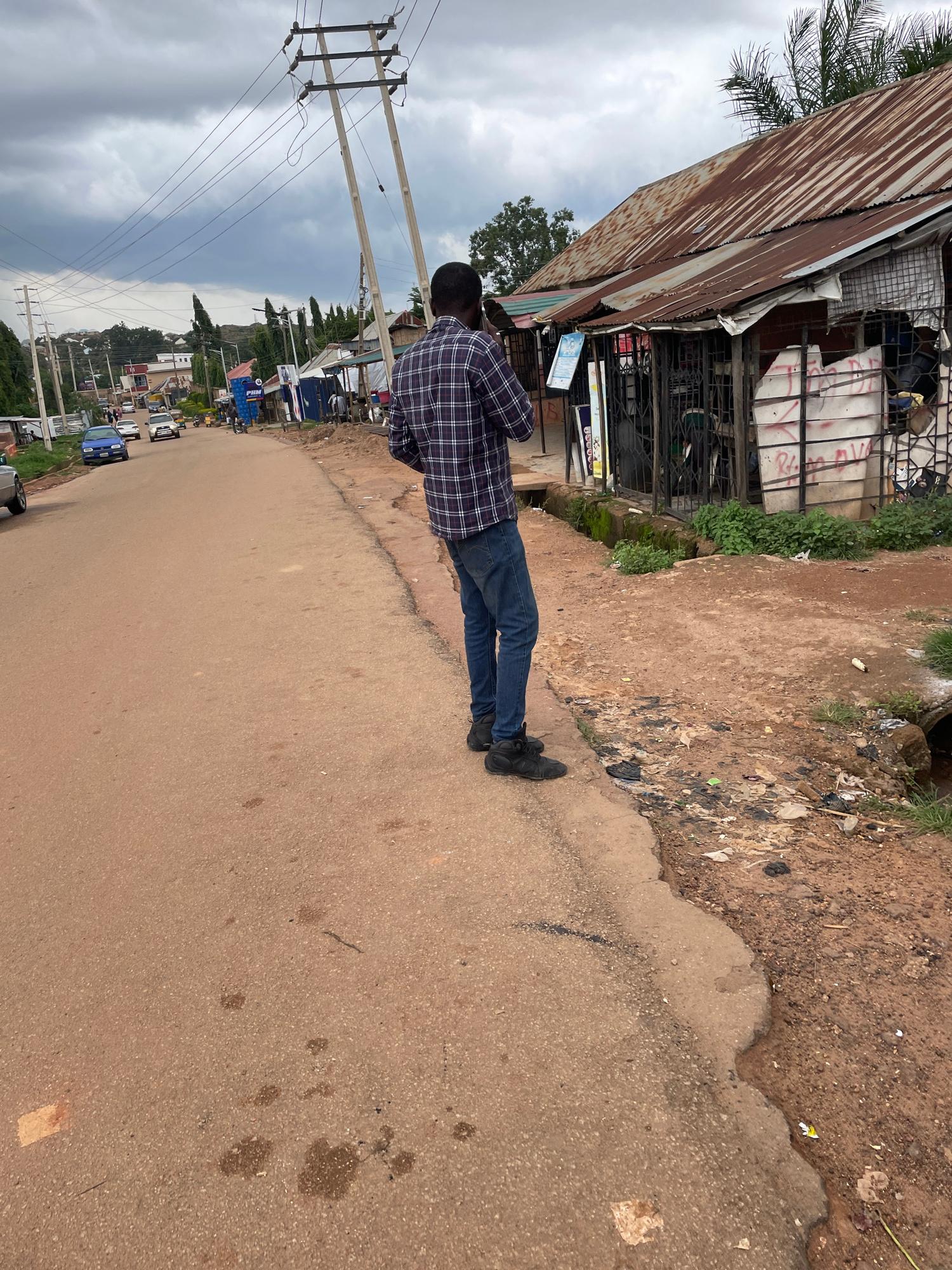 An over-the-shoulder, medium shot captures a person standing on the side of a dirt road, facing away from the camera. The person is wearing a plaid shirt and blue jeans and is holding a cell phone to their ear, appearing to be in a conversation. The environment is an urban setting, possibly in Jos, Nigeria, with a mix of dilapidated buildings and a busy street. A corrugated iron roof structure is visible to the right, with metal bars and graffiti on its facade. A narrow gutter runs along the side of the road, filled with debris and stagnant water. Utility poles with electrical wires stretch across the cloudy sky in the background. Cars and other vehicles are seen driving down the road, suggesting an active street scene. The ground is a mixture of red soil and asphalt, with scattered litter indicating a lack of proper waste management.