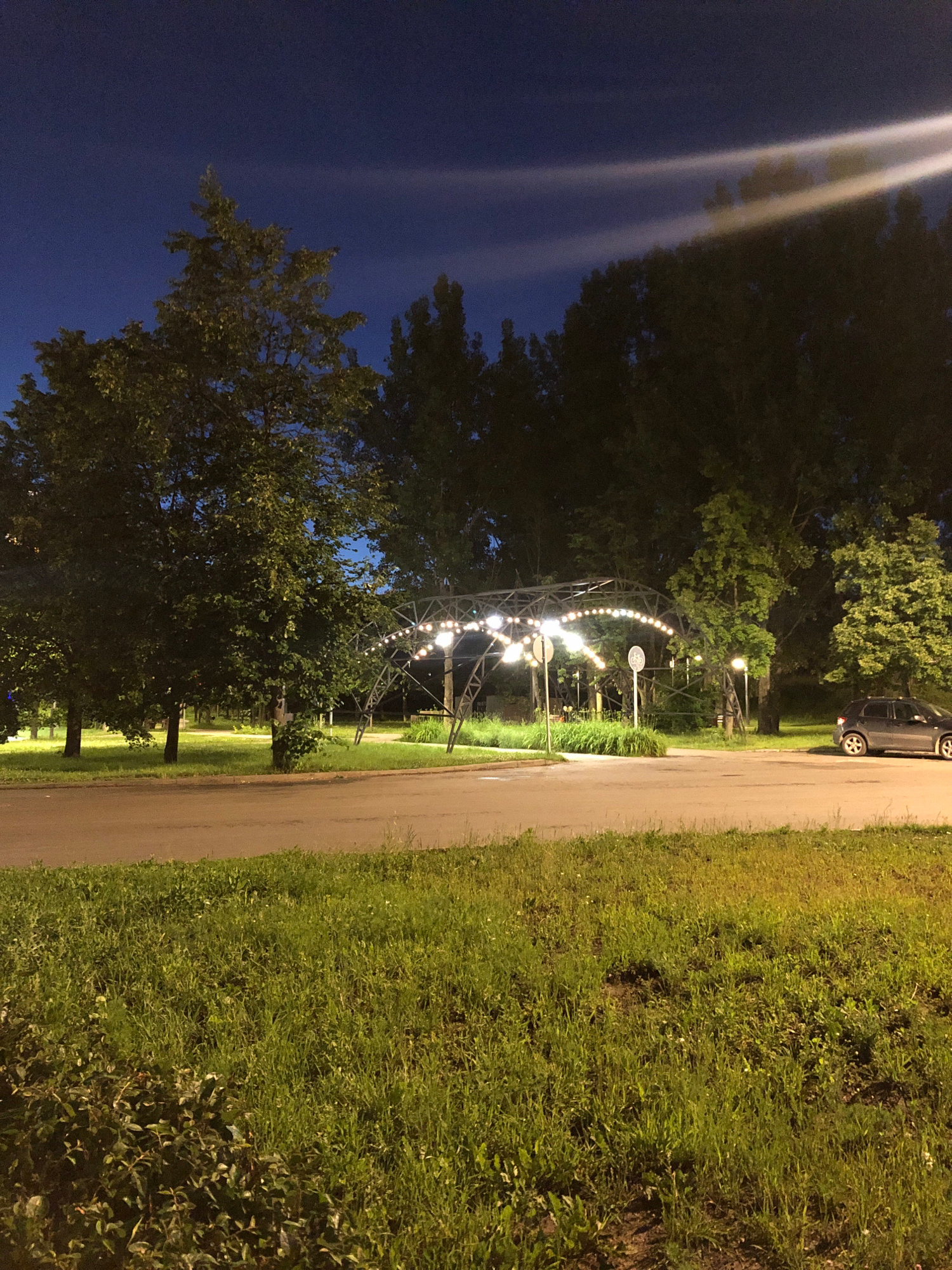 An elevated, arched structure adorned with string lights is visible in a park at night. The structure appears to be a canopy or pavilion, with a metal frame supporting numerous small, round lights. The park is characterized by lush green grass and several mature trees with dense foliage. A paved road runs across the foreground of the image, with a strip of grass separating it from the park area. On the right side of the road, a dark-colored car is parked. The sky above is a deep blue, indicating it is twilight or early night. The overall scene suggests an outdoor public space, possibly a recreational area or an event venue, illuminated by artificial lighting against the dark sky.