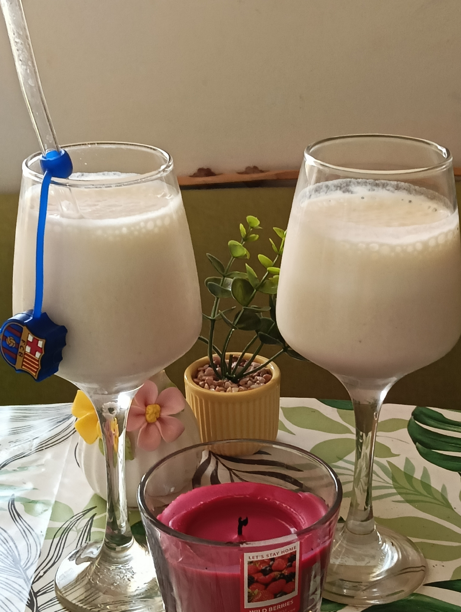 Two tall glasses of a creamy white beverage, possibly a milkshake or smoothie, are set on a table. A clear straw with a blue holder is in the left glass, adorned with a blue and red charm featuring a crest. The right glass has no straw visible. In the background, a small potted plant with green leaves sits in a yellow pot. A white ceramic decorative item with two fabric flowers, one pink and one yellow, is positioned behind the left glass. A red scented candle in a glass jar, labeled "LET'S STAY HOME WILD BERRIES," is in the foreground. The table is covered with a tablecloth featuring a pattern of green leaves. The lighting suggests daytime, and the overall scene evokes a relaxed and possibly domestic setting. The location context provided is Harmanli, Bulgaria.