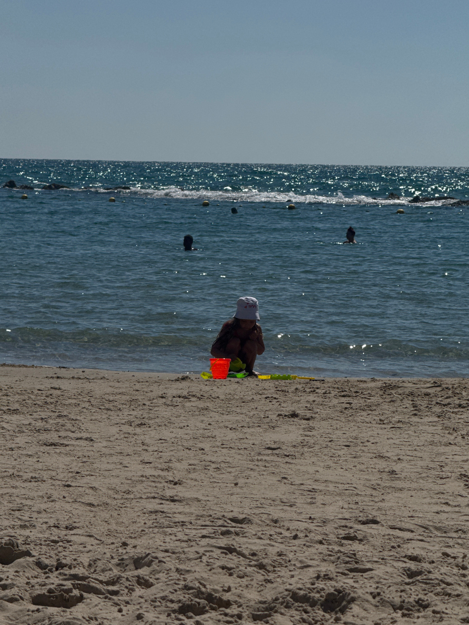 This is a vertical, eye-level photograph taken on a beach. The scene depicts a young child playing in the sand near the water's edge. The child is wearing a white hat and is partially visible from the back. They are engaged in an activity with a bright orange bucket and some green plastic toys.
The beach has a sandy shore with visible texture, and the ocean water is calm and blue, with small waves lapping at the sand. There are several dark, spherical buoys visible in the water, indicating a possible marked-off swimming area or a net. The sunlight reflects brightly off the water's surface, suggesting it is daytime, likely sunny. The overall atmosphere appears peaceful and relaxed, typical of a beach day. The image does not contain any visible text.