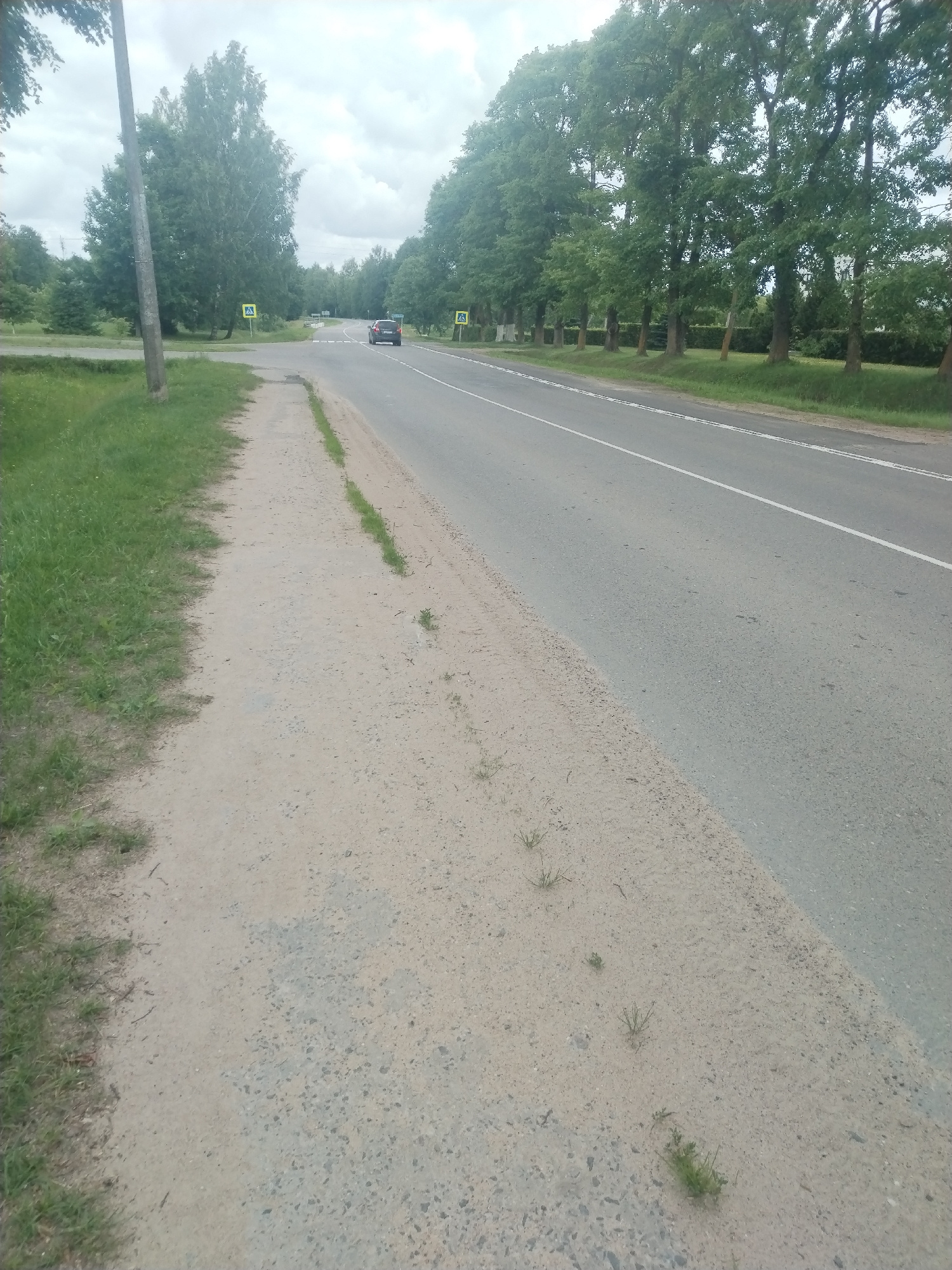 This media file depicts a paved road curving to the right, with a sandy shoulder on the left side and a grassy verge beyond that. Tall trees line the right side of the road, forming a dense canopy. A single lane of traffic is visible, with a white line marking the center. In the distance, a red car is traveling away from the viewer. Two yellow pedestrian crossing signs are visible on the left side of the road, suggesting a populated area or a road with pedestrian traffic. The sky is overcast, indicating a cloudy or possibly rainy day. The overall setting appears to be rural or suburban, with natural elements like trees and grass contrasting with the man-made road. There are no people clearly visible in the image. The time of day is indeterminate due to the overcast sky, but it appears to be daytime. The location context is Pastavy, Belarus.