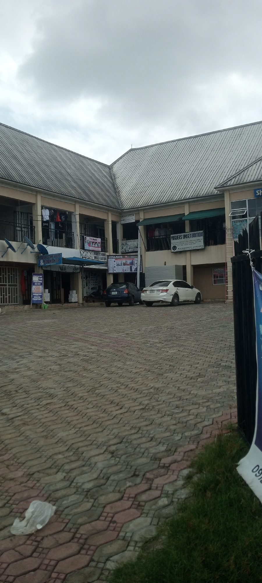 The image captures a commercial building with multiple shops on the ground floor and balconies on the upper floor, situated in Jalingo, Nigeria. The building features a corrugated metal roof and is constructed with beige-colored walls. Paved brickwork forms the ground in front of the building, where two cars are parked: a dark blue sedan and a white sedan. Several signs are visible, advertising businesses such as "YOGUENS UNISEX BOUTIQUE," "MEGA WAVE," and "SOPHIEZ." A blue awning is extended over one of the shops. The sky is overcast with gray clouds. A plastic bag is lying on the ground in the foreground.