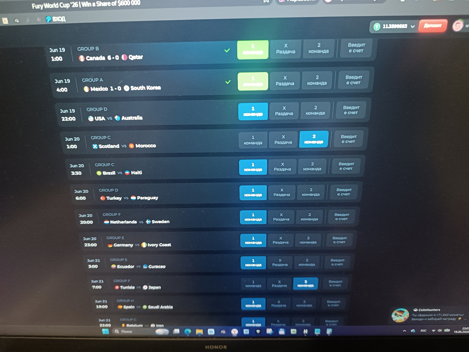 The image shows a computer screen displaying a betting or gaming interface for a "Fury World Cup '26". The screen shows a list of upcoming soccer matches, grouped by tournament stages. Each match listing includes the date and time, participating teams with their national flags, and betting options labeled as "1 команда" (Team 1), "X Раздача" (Draw), and "2 команда" (Team 2). Some matches have checkmarks next to them, indicating a selection has been made. At the top right of the screen, a balance of "11.35998583" is displayed, along with a "Deposit" button. At the bottom right, there is a notification from "CoinHunters" about accumulating coins and claiming a prize. The interface language appears to be Russian. The image is taken from an angle, suggesting it's a photograph of a monitor. There is no visible indication or information within the image that relates to the city of Simferopol, Ukraine.