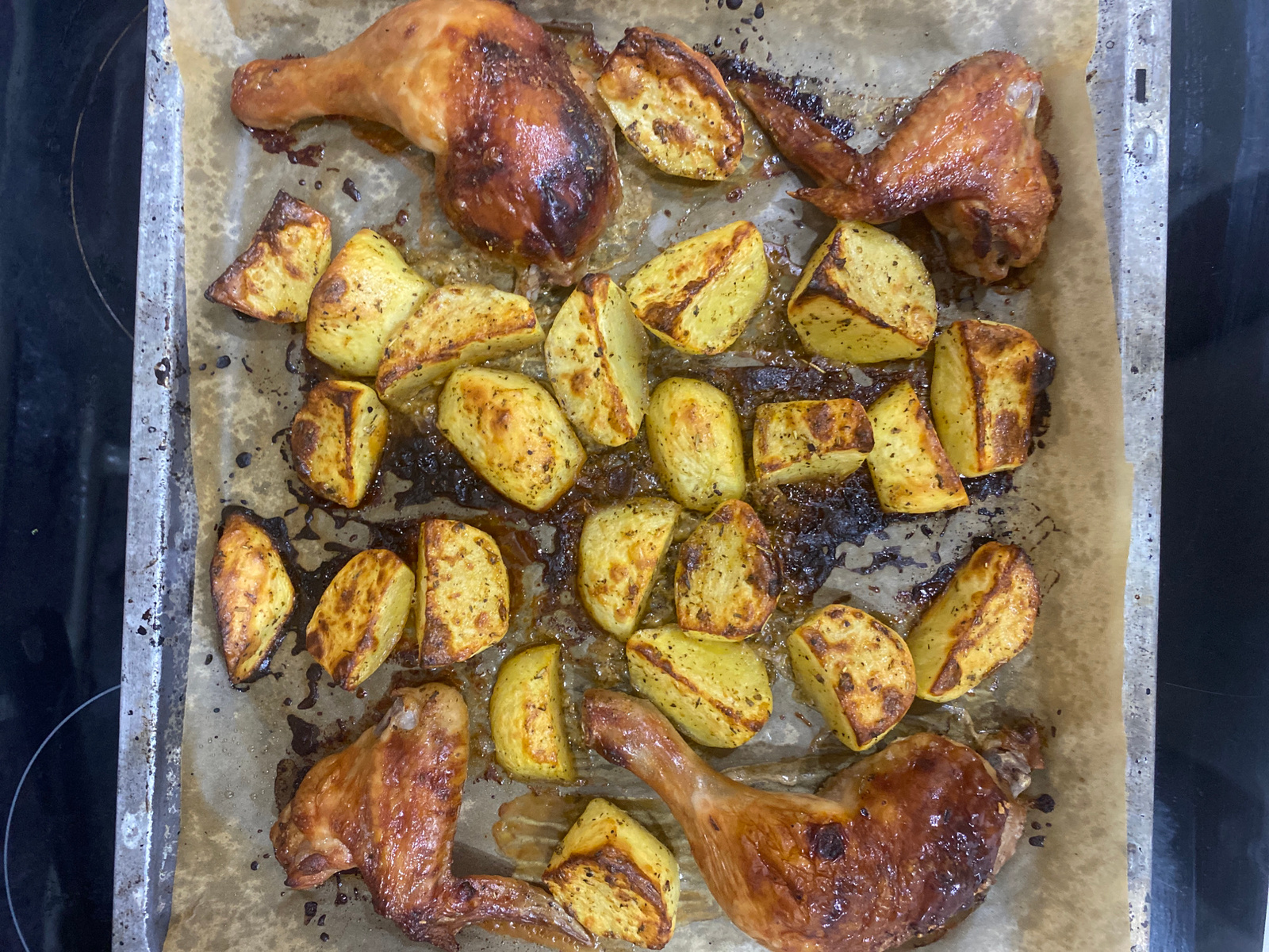 A top-down, eye-level, medium shot captures a baking sheet filled with roasted chicken and potato wedges. The baking sheet, lined with parchment paper, rests on a dark stovetop, likely in a kitchen. The chicken pieces, a mix of legs and wings, are golden brown and appear juicy with some crispy skin. The potato wedges are also golden brown, some showing slight charring, and are seasoned with visible herbs. The parchment paper has some dark, greasy spots from the cooking process. The background is dominated by the dark surface of the stovetop. There is no indication of the location being Novokuybyshevsk, Russia, or any other specific city. The image focuses solely on the food.