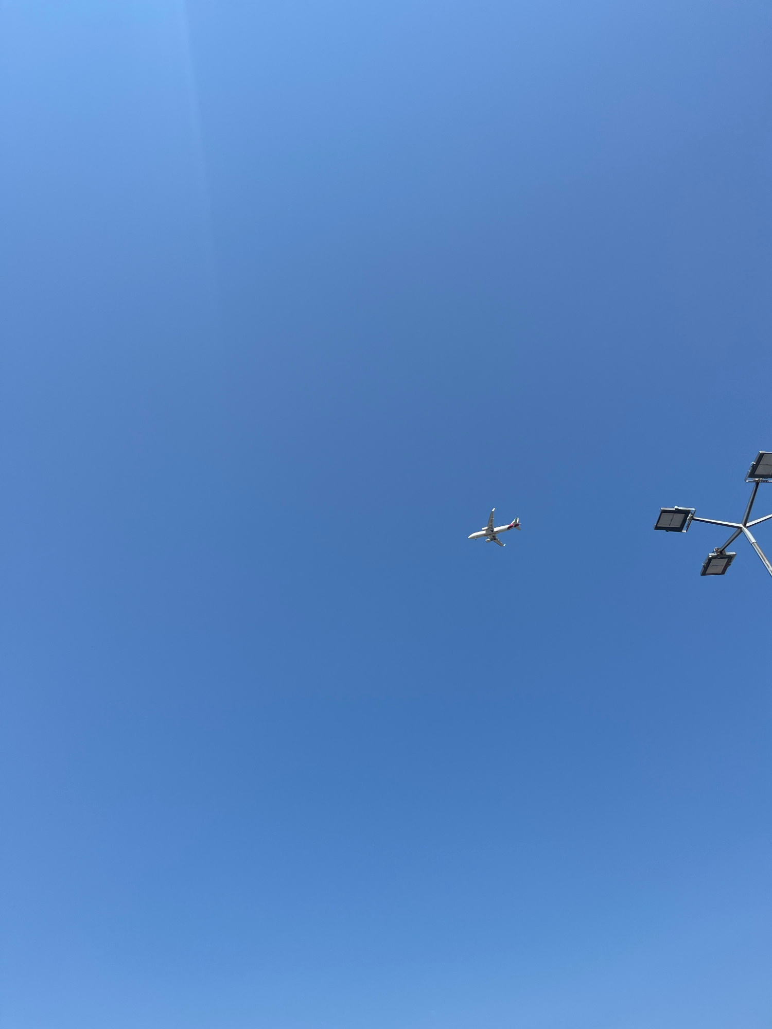 An airplane is captured mid-flight against a clear blue sky. To the right of the frame, a portion of a modern street lamp with four square lighting fixtures is visible. The scene appears to be an outdoor public space in Piaseczno, Poland. There are no people or other discernible subjects in the image.