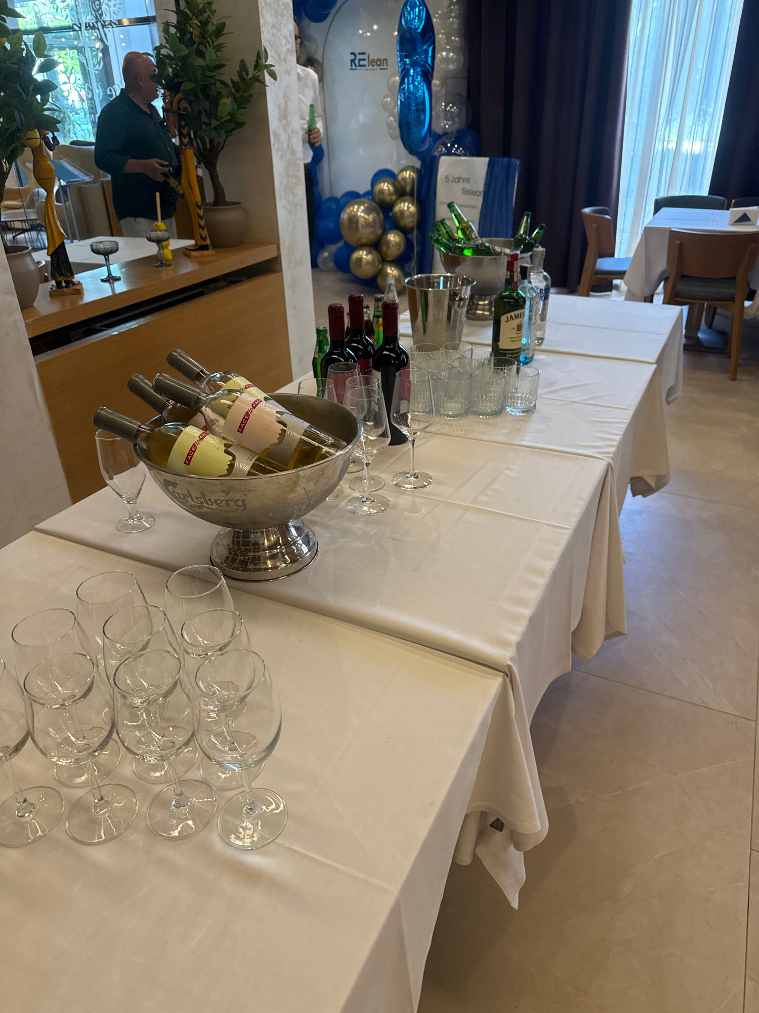 The image depicts a table set for an event, likely a reception or celebration, with a focus on alcoholic beverages and glassware. The table is covered with a white tablecloth and displays several bottles of wine, a silver ice bucket filled with bottles of what appears to be beer, and a variety of wine glasses and tumblers. Among the bottles, a green bottle of Jameson Irish Whiskey is visible. In the background, balloons are arranged, suggesting a festive atmosphere. A man in a dark shirt is standing near a mirrored surface, and another person, partially visible, is holding a green bottle. The setting appears to be indoors, possibly a restaurant or banquet hall, with natural light coming from windows covered by curtains. The floor is tiled. The scene is in Sofia, Bulgaria, as indicated by the context provided.