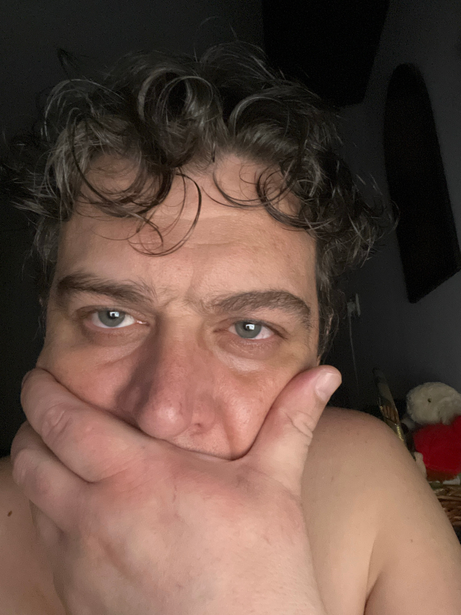 A close-up, low-angle, selfie-style photograph shows a person's face and upper chest, cropped from the shoulders up, against a dark, indistinct background. The individual has curly, dark brown hair with some graying strands, and is looking directly at the camera with a contemplative or concerned expression. Their right hand is raised to their face, covering their mouth and nose, with their thumb resting on their cheekbone. Their eyes are visible, appearing to be a light green or gray color. The skin tone is fair, with some redness on the cheeks and around the eyes. The background is dark, with a faint shadow on the right side of the frame that suggests an arched opening or corner. A small, out-of-focus object, possibly a stuffed toy with a red piece of fabric, is visible in the lower right corner, suggesting an indoor setting. The location in Almaty, Kazakhstan, cannot be determined from the visual information provided.