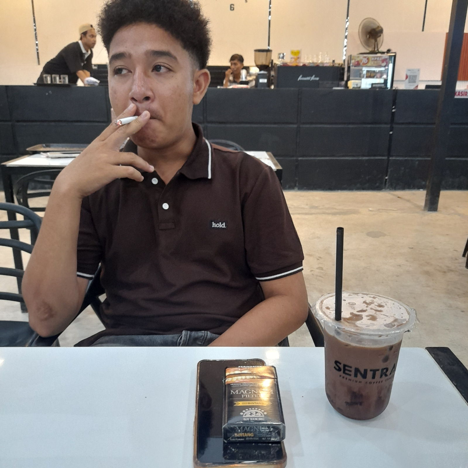 The image shows a young man smoking a cigarette while sitting at a table in what appears to be a cafe in Lhokseumawe, Indonesia. He is wearing a brown polo shirt. On the table in front of him is a smartphone with a pack of "MAGNA FILTER" cigarettes placed on top of it, and a cup of iced coffee from "SENTRA PREMIUM COFFEE". In the background, other people are visible, including a barista behind a counter. The cafe has black walls and a light-colored floor.