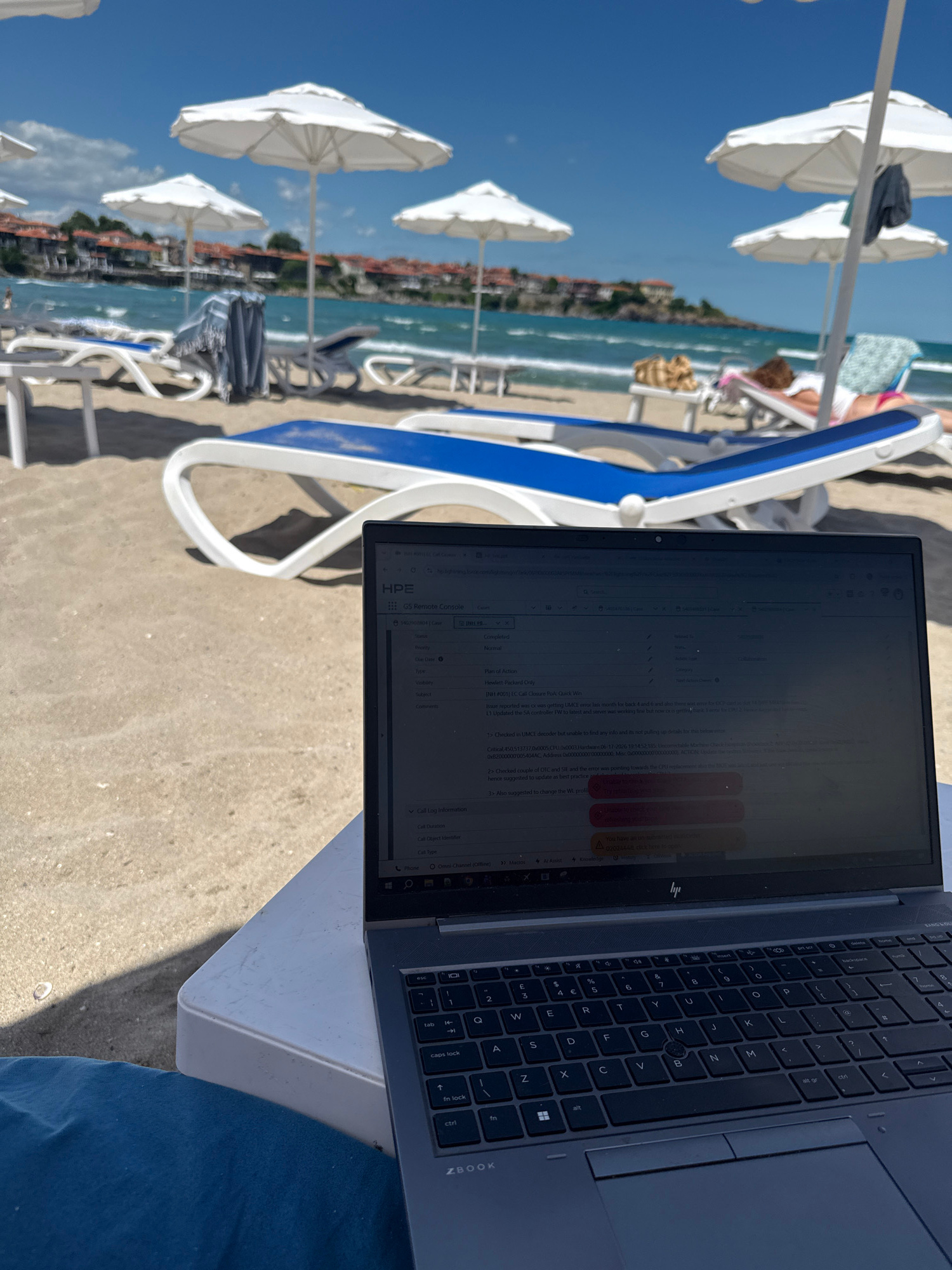 A laptop is positioned on a white table, open and displaying a software interface. The laptop screen shows a grid-like structure with text and colored boxes, suggesting a work-related application. The scene is set on a beach, with sand visible in the foreground and around the table. In the background, lounge chairs and white umbrellas are arranged along the coastline, with the sea and a distant town visible beyond. The town, with its red-tiled roofs, is identifiable as Sozopol, Bulgaria, a historic coastal city. The sky is blue with some clouds, indicating a sunny day.