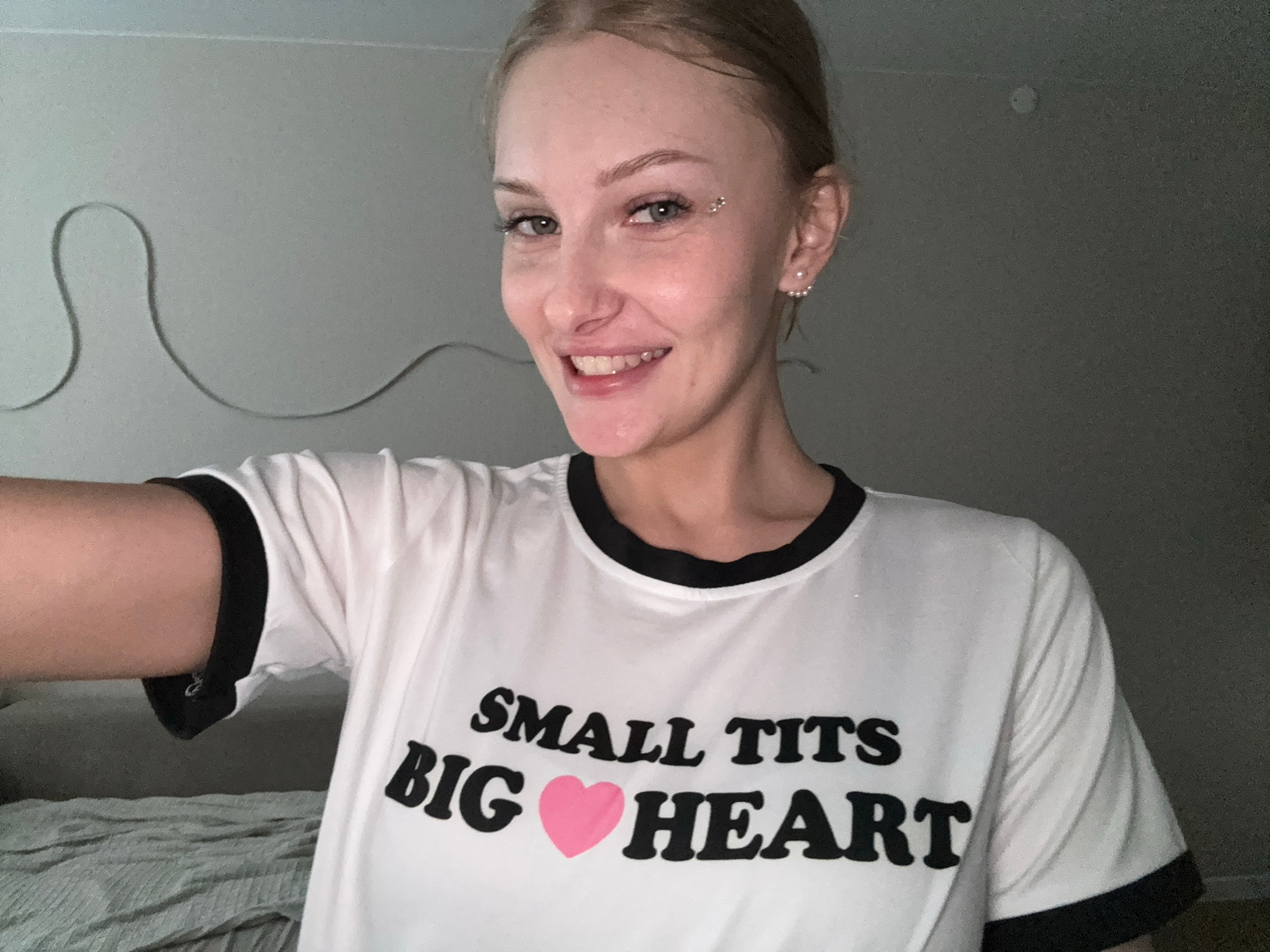 The image shows a young woman with blonde hair and blue eyes, smiling at the camera. She is wearing a white t-shirt with black trim and a graphic that reads "SMALL TITS BIG HEART" with a pink heart symbol. She has piercings on her face and ears. The background appears to be a room with a wall and a bed. There are no discernible features that would indicate the location is the city of Perm, Russia.
