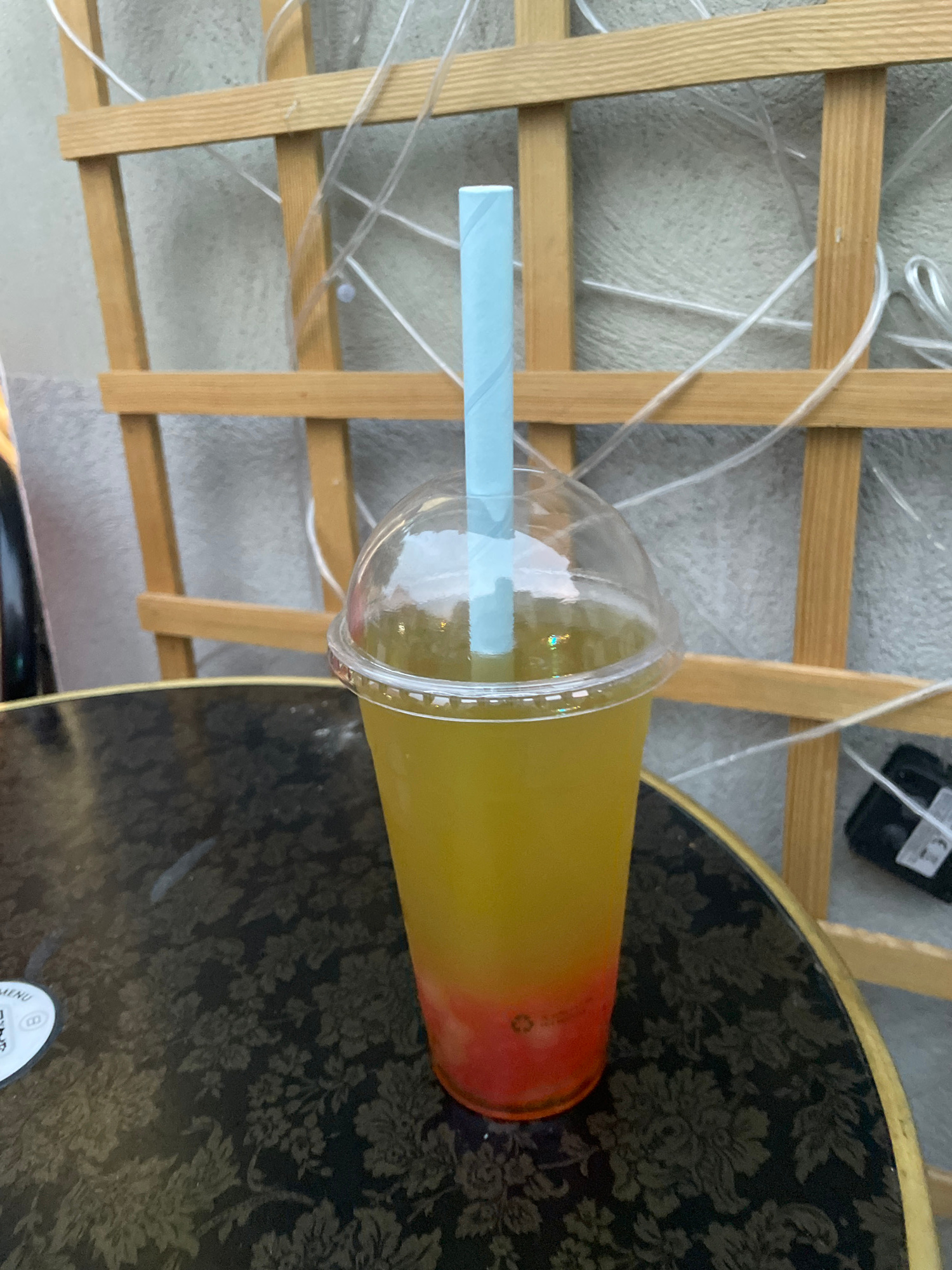 A single plastic cup filled with a layered beverage sits on a dark, patterned table. The beverage is predominantly yellow, with a bright red layer at the bottom. A light blue straw extends from the translucent lid. In the background, a wooden trellis with clear plastic tubing crisscrosses it. The scene appears to be outdoors, with a textured wall behind the trellis. A small white circular object with text and a symbol is partially visible on the table to the left of the cup.