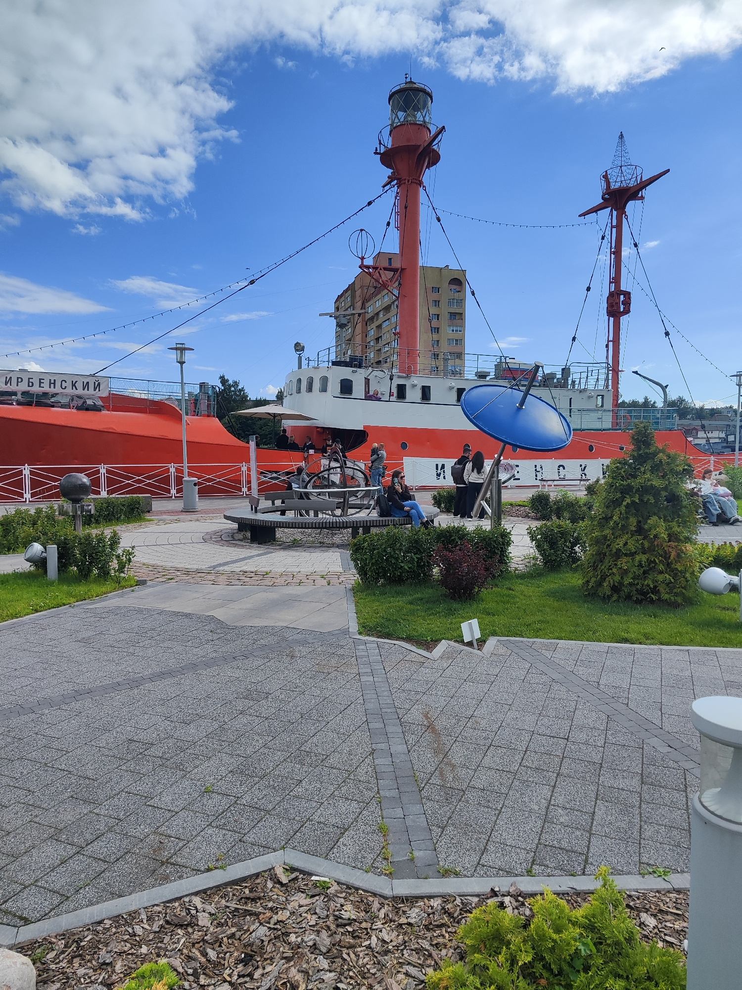 This image shows the "Irıbenskıi" lighthouse ship, a former vessel that has been converted into a museum and is docked in Kaliningrad, Russia. The ship is predominantly orange and white, with a tall red lighthouse structure at the front. In the foreground, a paved walkway leads towards the ship, with benches and greenery along the sides. Several people are seated on the benches, appearing to be visitors. A large blue satellite dish is mounted on a pole near the ship. The sky is blue with scattered white clouds. The overall scene depicts a public space with a historical maritime attraction.