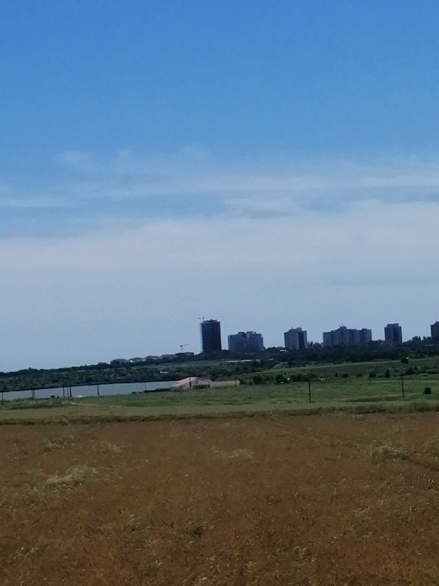 The image shows a distant cityscape with several tall buildings under construction, set against a bright blue sky with scattered clouds. The foreground is dominated by a dry, golden-brown field of grass or crops. In the middle ground, a body of water is visible, bordered by a line of trees and the construction site. A small, light-colored structure with a reddish roof is situated near the water. The overall scene suggests an urban development project in a semi-rural or suburban setting.