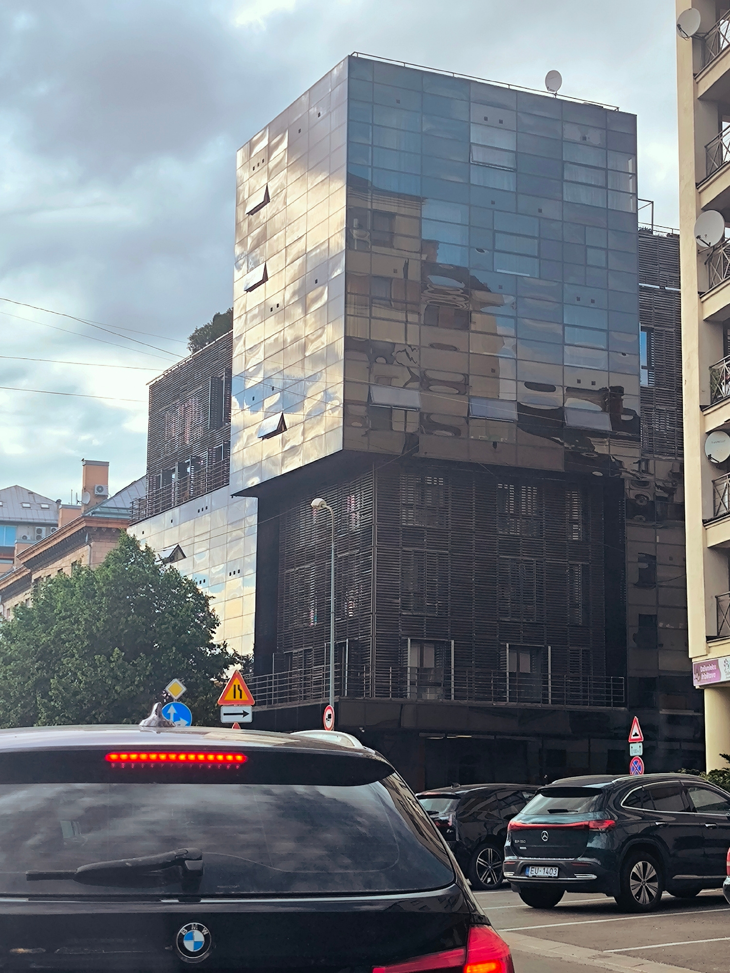 This is a slightly elevated, medium shot captured from behind a dark-colored SUV on a cloudy day in Riga, Latvia. The SUV's brake lights are illuminated red. To the right of the SUV, a dark teal Mercedes-Benz EQE with a European license plate EU-1403 is parked. In the background, a modern, multi-story building with a reflective glass facade dominates the scene. The building has several open windows and features horizontal louvers on lower floors. Adjacent to this building, to the right, is a lighter-colored building with balconies. Traffic signs are visible on the left, including directional arrows and a warning sign. Trees and other buildings are present further in the background. The sky is overcast with a significant amount of cloud cover.