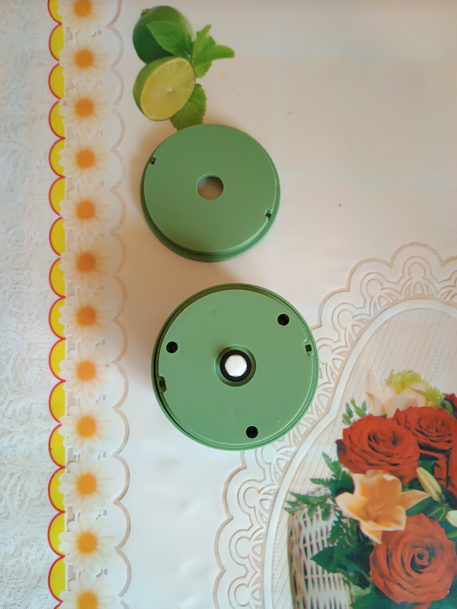 The image shows two green circular objects with a hole in the center of the top one and a white circular fixture in the center of the bottom one. The objects are placed on a white surface with a floral pattern. To the left, there is a border of daisies with yellow centers and red and yellow circular accents. To the upper left, there are two lime halves and some green leaves. To the right, there is a decorative white lace pattern and a basket of red and orange flowers. The scene is indoors and appears to be a flat surface, possibly a table or countertop. There is no visible indication of the city of Zaporizhzhia, Ukraine.
